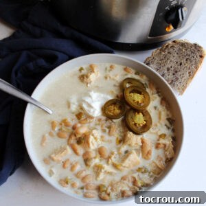 Crockpot White Chicken Chili Bowl of white chicken chili served with sour cream, jalapeno slices and sourdough bread in front of slow cooker it was cooked in.