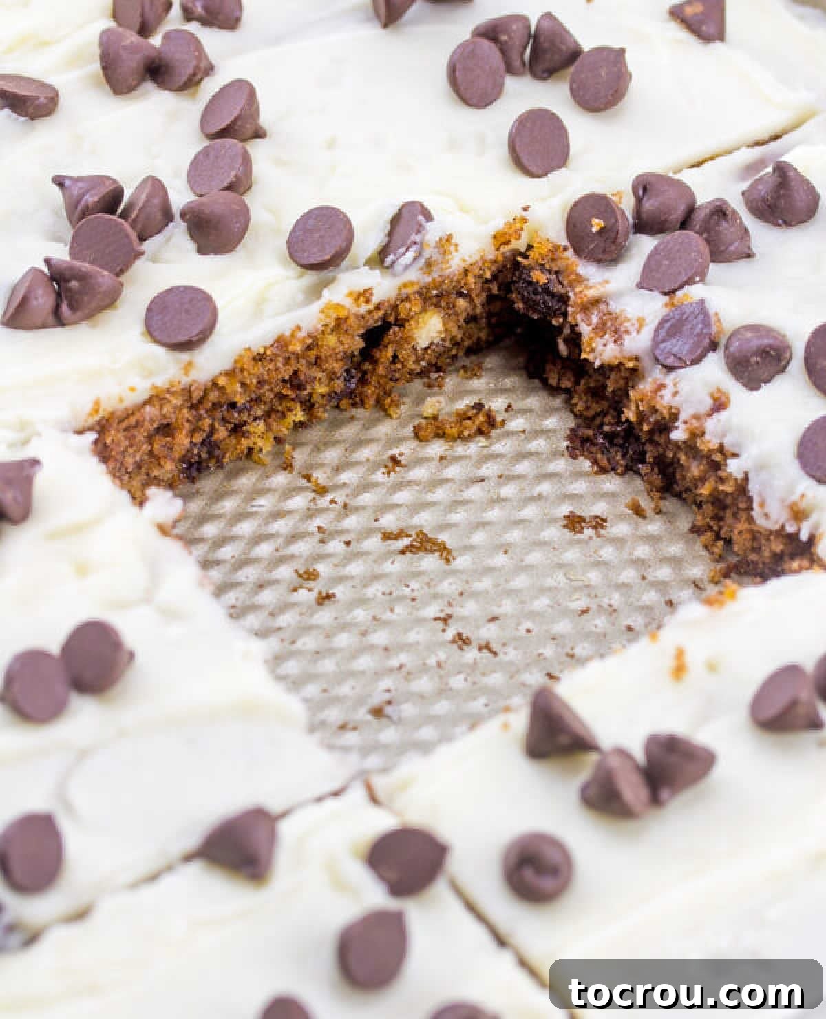 The Ultimate Chocolate Chip Cookie Dough Texas Sheet Cake 4 sheet pan filled with white icing topped chocolate chip Texas sheet cake with square piece missing, showing brown sugar cake topped with vanilla icing and chocolate chips.