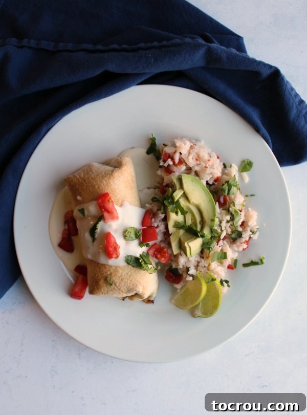 Crispy Baked Barbacoa Chimichangas 5 Overhead view of a dinner plate featuring a baked chimichanga smothered in queso, accompanied by Mexican rice, fresh avocado slices, and lime wedges.