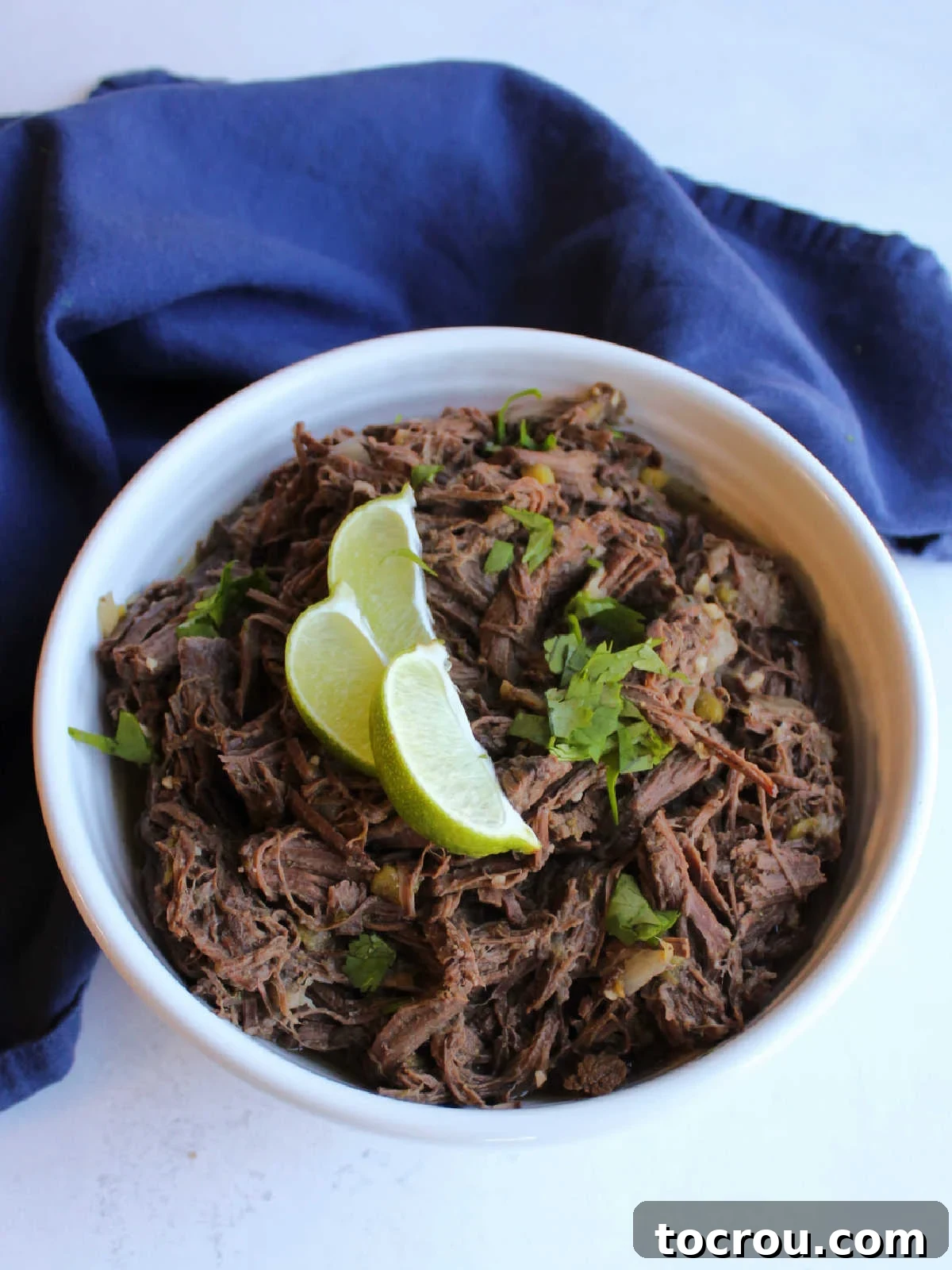 Serving bowl filled with shredded venison barbacoa, garnished with fresh chopped cilantro and bright lime wedges.