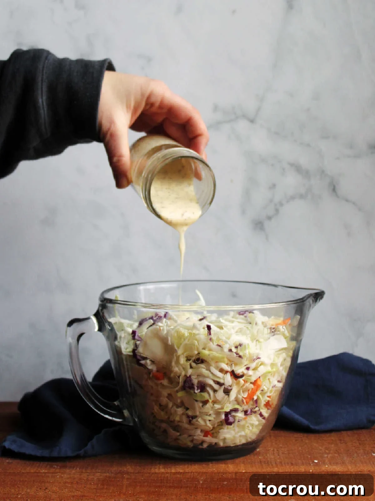 Pouring the Dressing A hand pouring homemade condensed milk coleslaw dressing over shredded cabbage in a bowl.