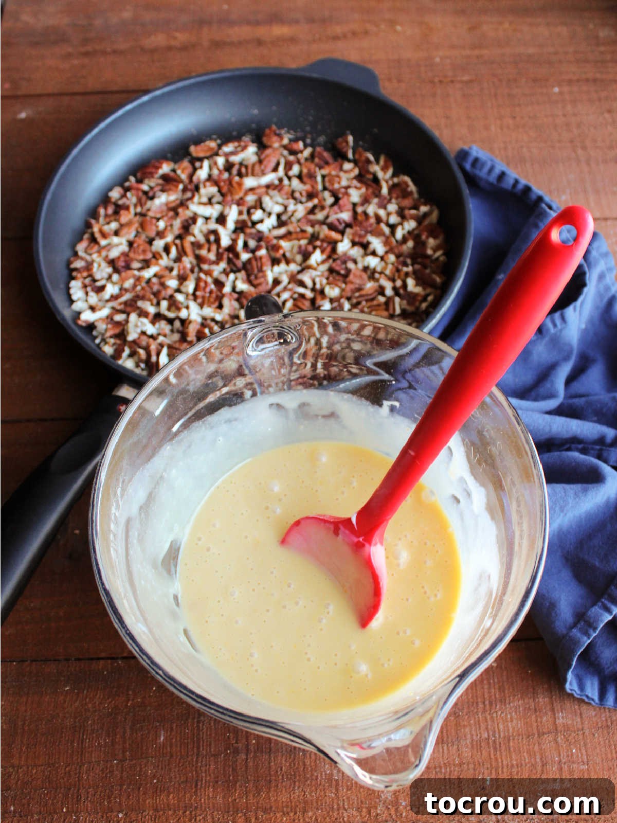 Mixing bowl with condensed milk pie filling inside next to skillet with toasted pecans.
