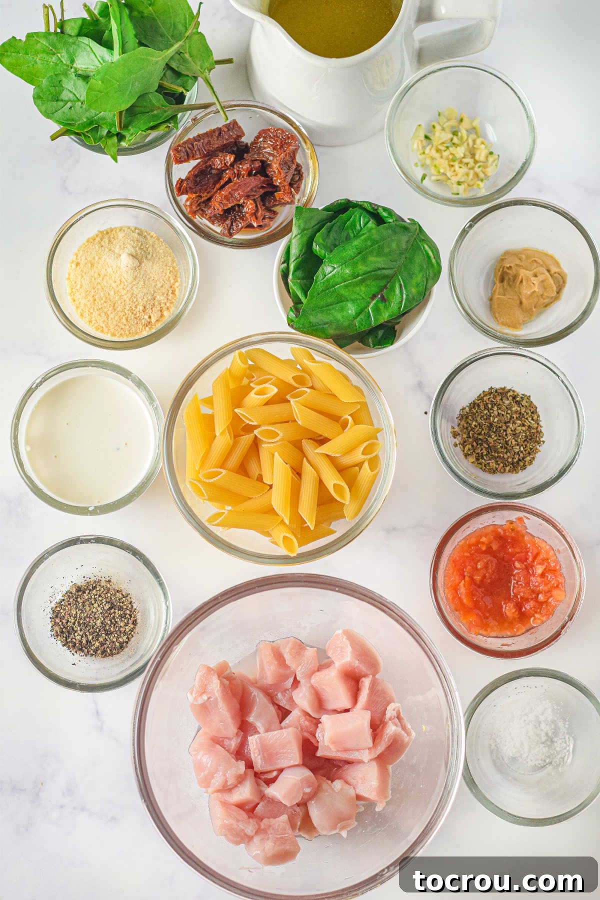 Ingredients for creamy Tuscan chicken pasta laid out: chicken, penne pasta, fresh spinach, cheese, sun-dried tomatoes, heavy cream, and various spices.