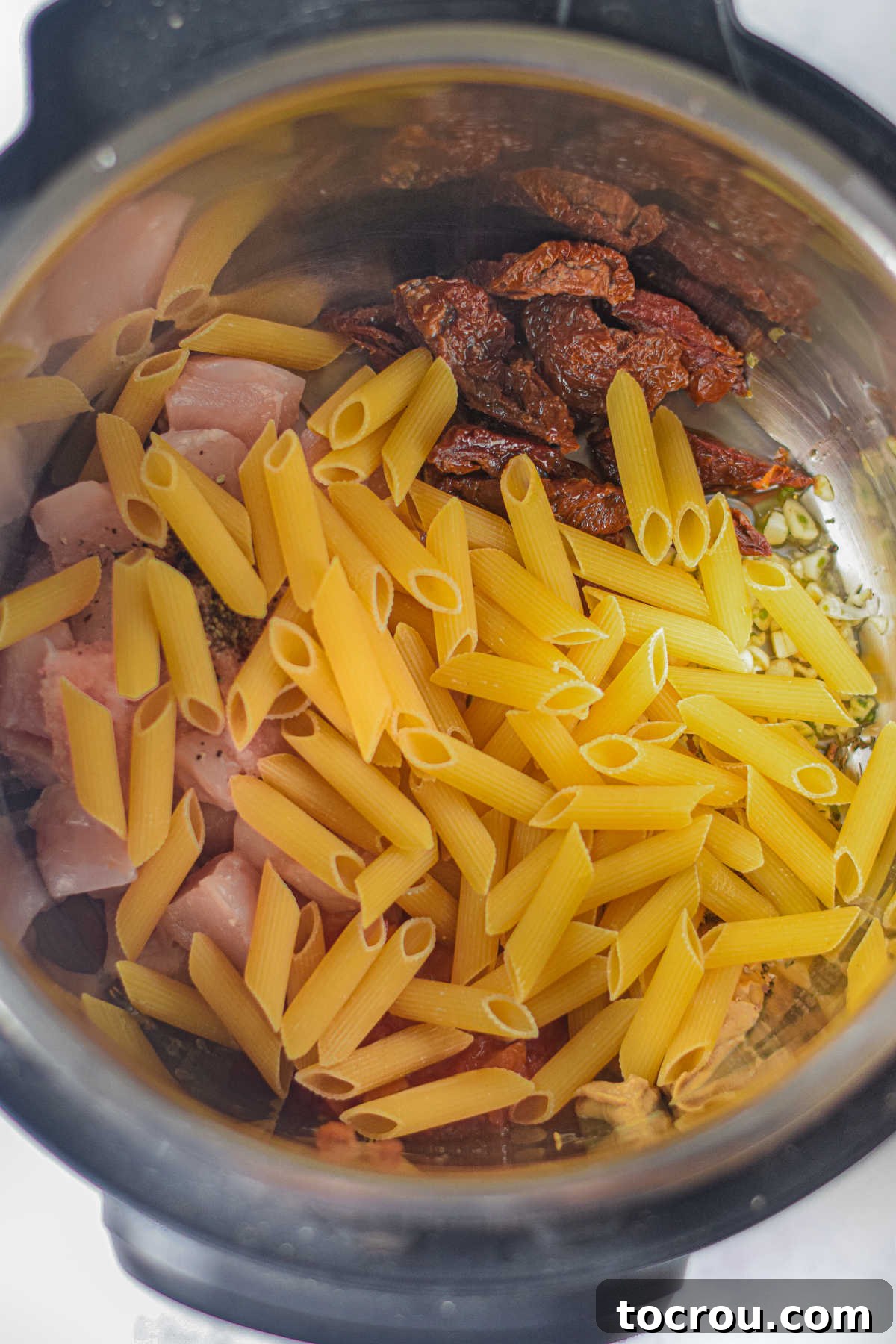 Uncooked penne pasta added on top of the chicken and seasonings in the Instant Pot, ready for pressure cooking.