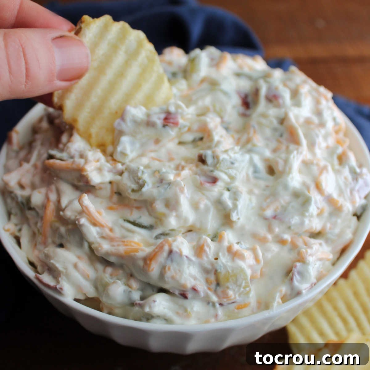 Dipping ruffled chip into creamy dill pickle dip with visible shredded cheddar cheese and dried beef