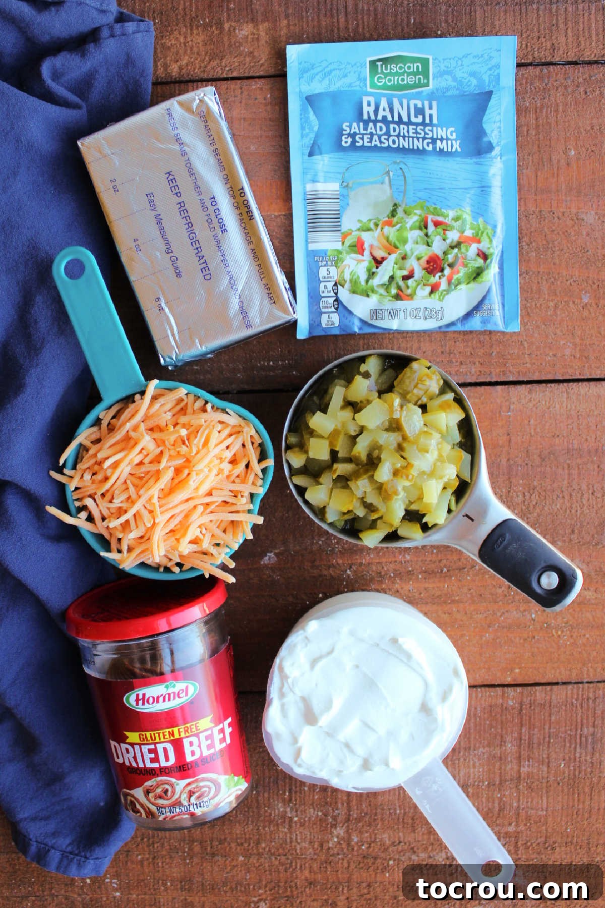 Assortment of ingredients for dill pickle dip: cream cheese, sour cream, ranch seasoning packet, a jar of dill pickles, shredded cheddar cheese, and dried beef