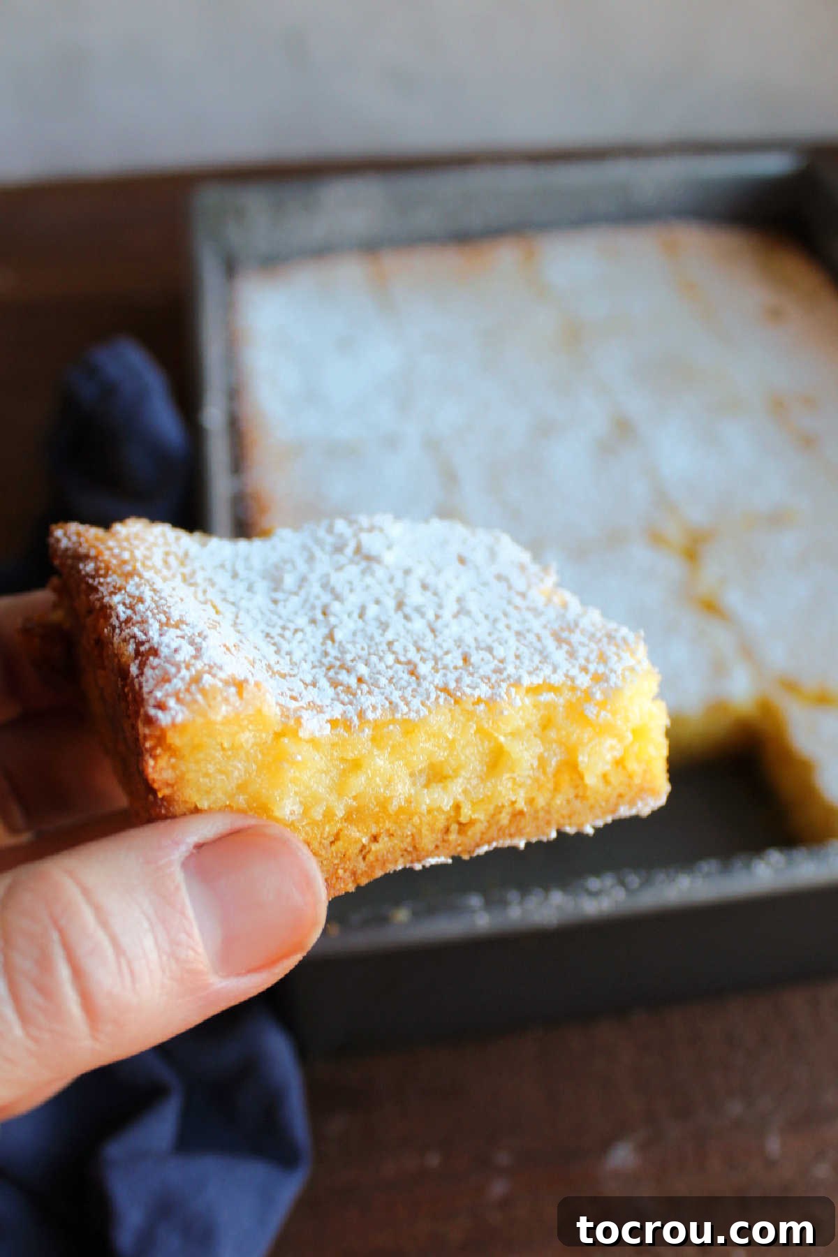 Zesty Lemon Chess Bars 3 Hand holding lemon gooey butter cake showing golden edge and soft gooey center.