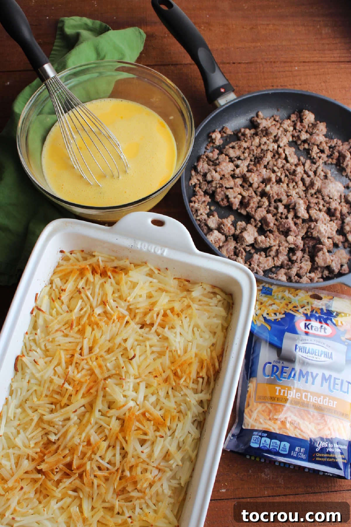 Casserole Ingredients Ready for Assembly A baking dish filled with partially cooked golden hashbrowns, alongside bowls of browned breakfast sausage, a whisked egg and milk mixture, and shredded cheddar cheese, all prepped for assembly into a delicious casserole.