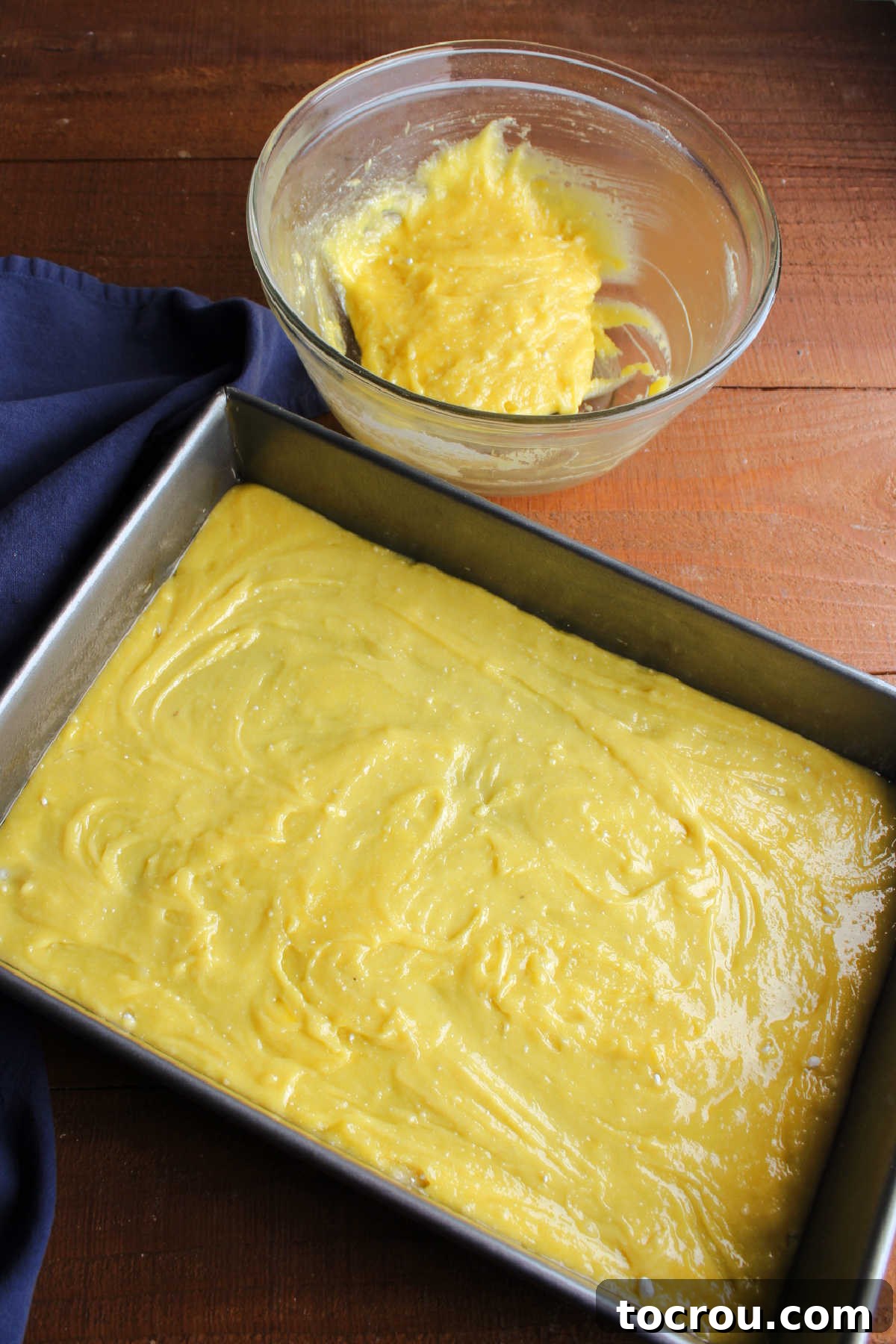 Cake Mix Crust in Baking Pan Two-thirds of the mixed cake mix dough pressed firmly into the bottom of a 9x13 inch baking pan, forming the crust. A small amount of dough remains in a white mixing bowl beside it.