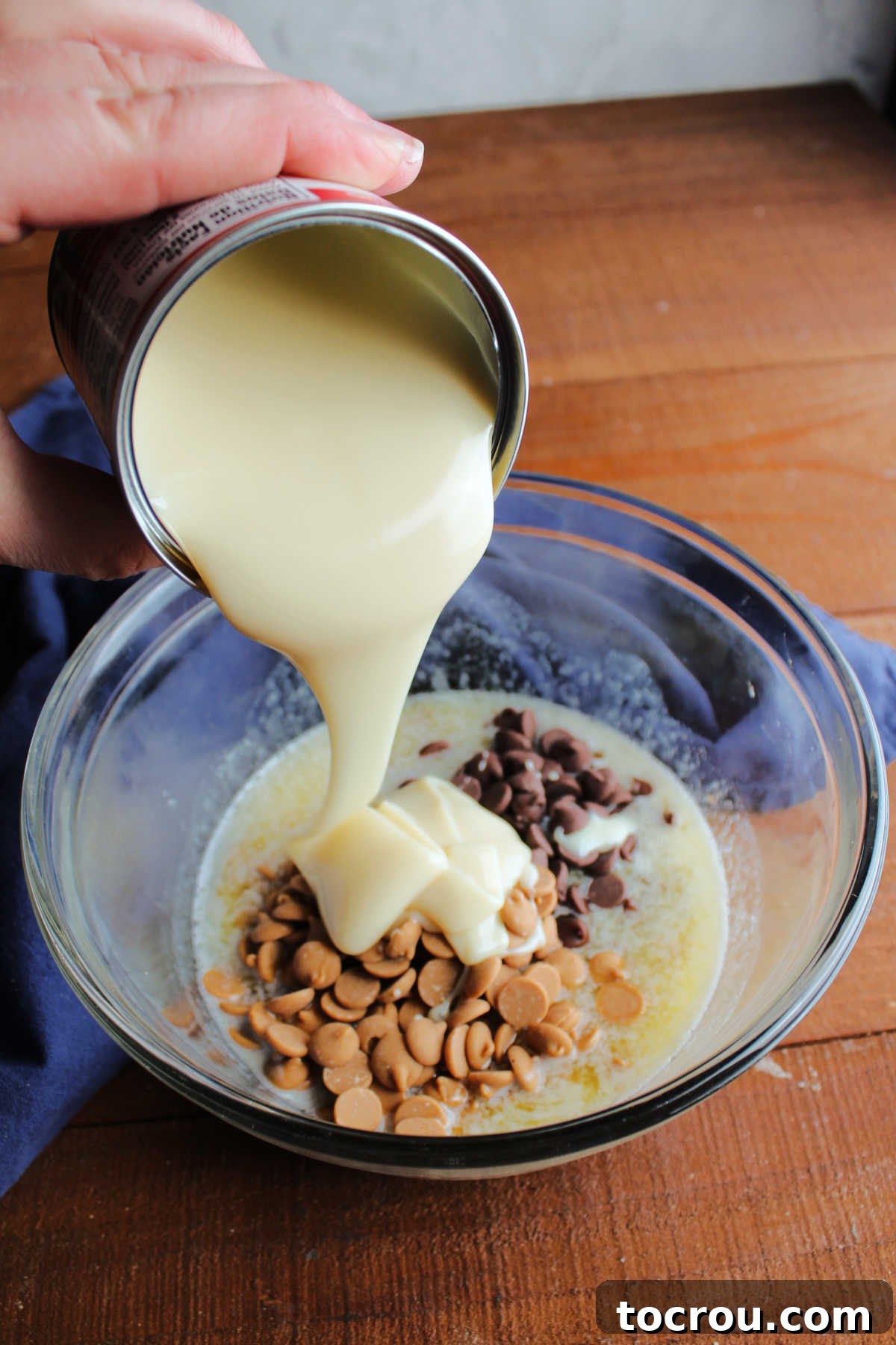 Adding Condensed Milk to Fudge Mixture Sweetened condensed milk being poured from a can into a glass mixing bowl, joining a mixture of melted butter, chocolate chips, and peanut butter chips.
