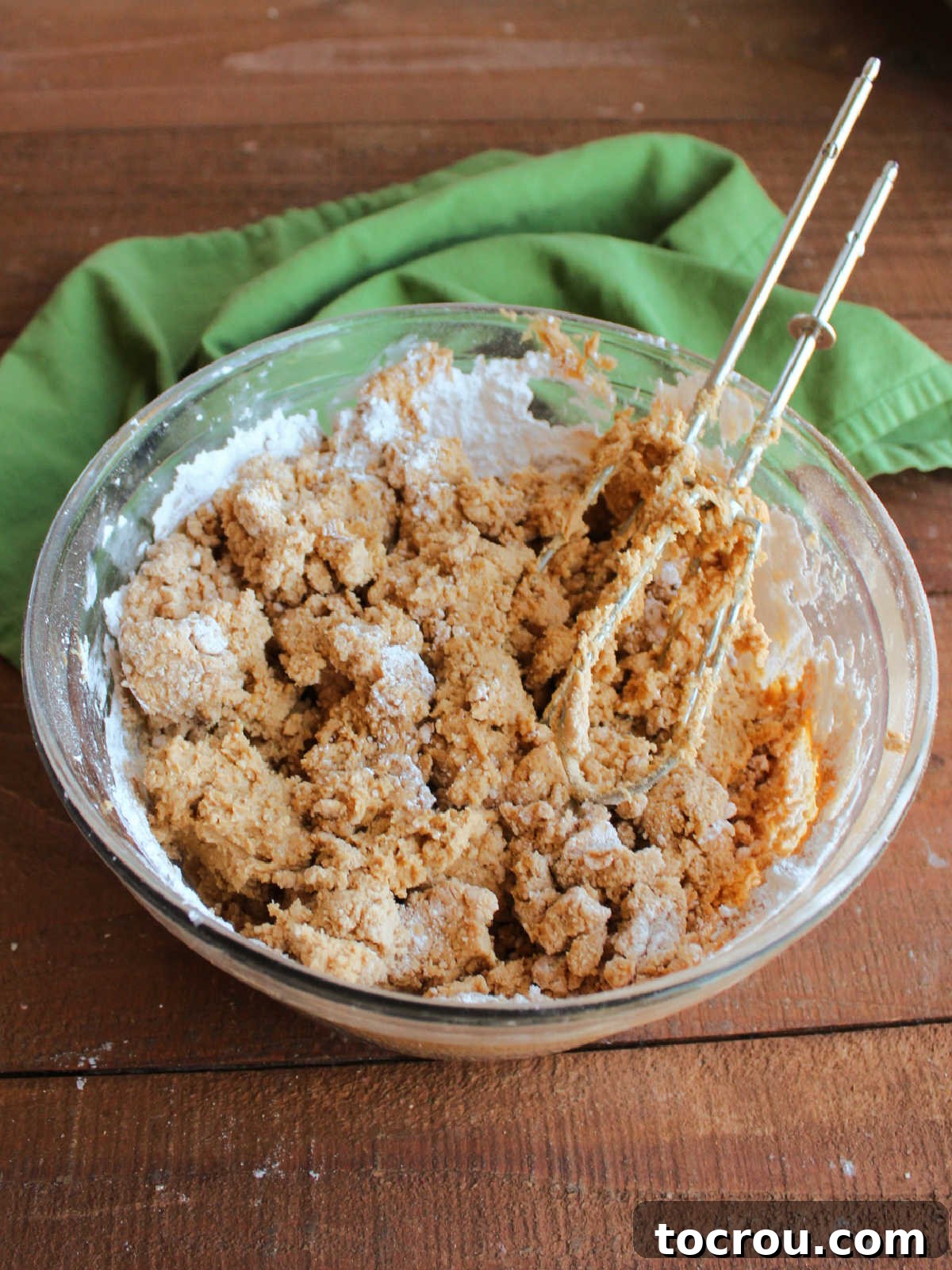 A bowl filled with a crumbly mixture of peanut butter and powdered sugar, with electric hand mixer beaters resting inside, showing the initial texture of the candy base.