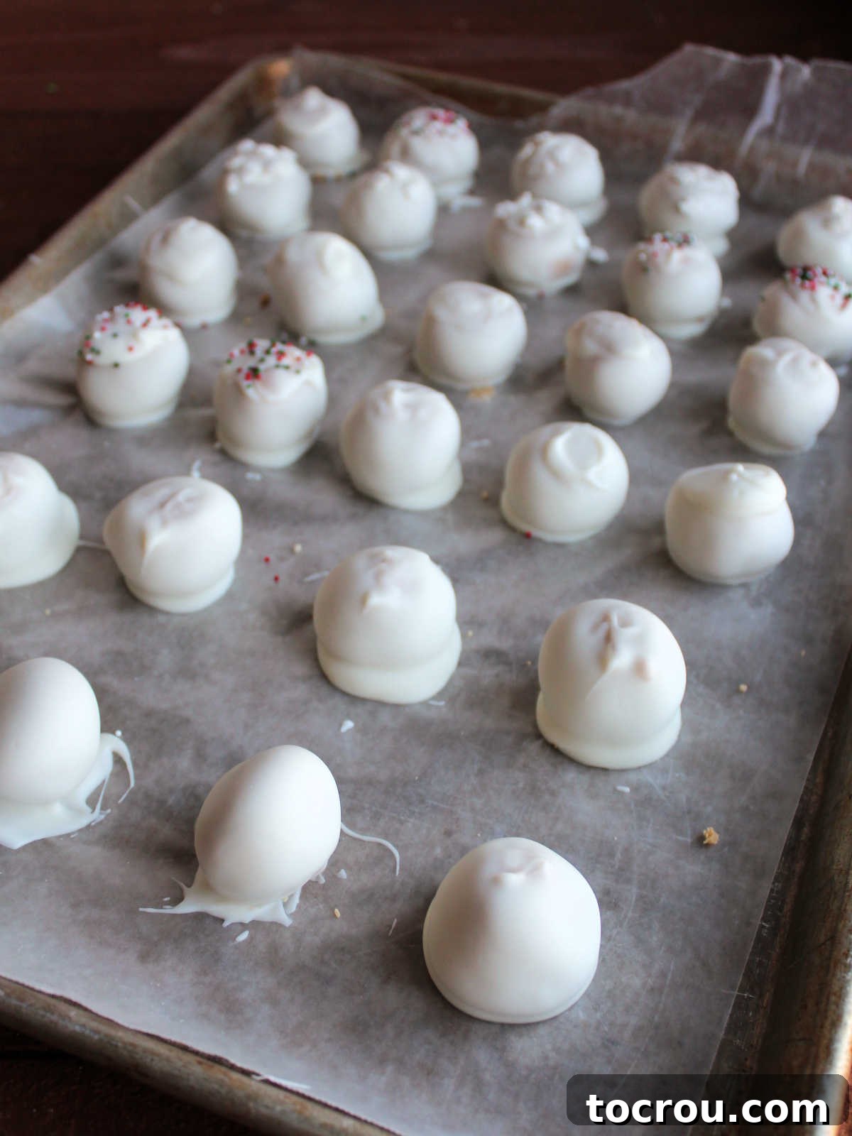 A baking tray filled with numerous white chocolate peanut butter snowballs, freshly and completely dipped in white chocolate, setting on wax paper.