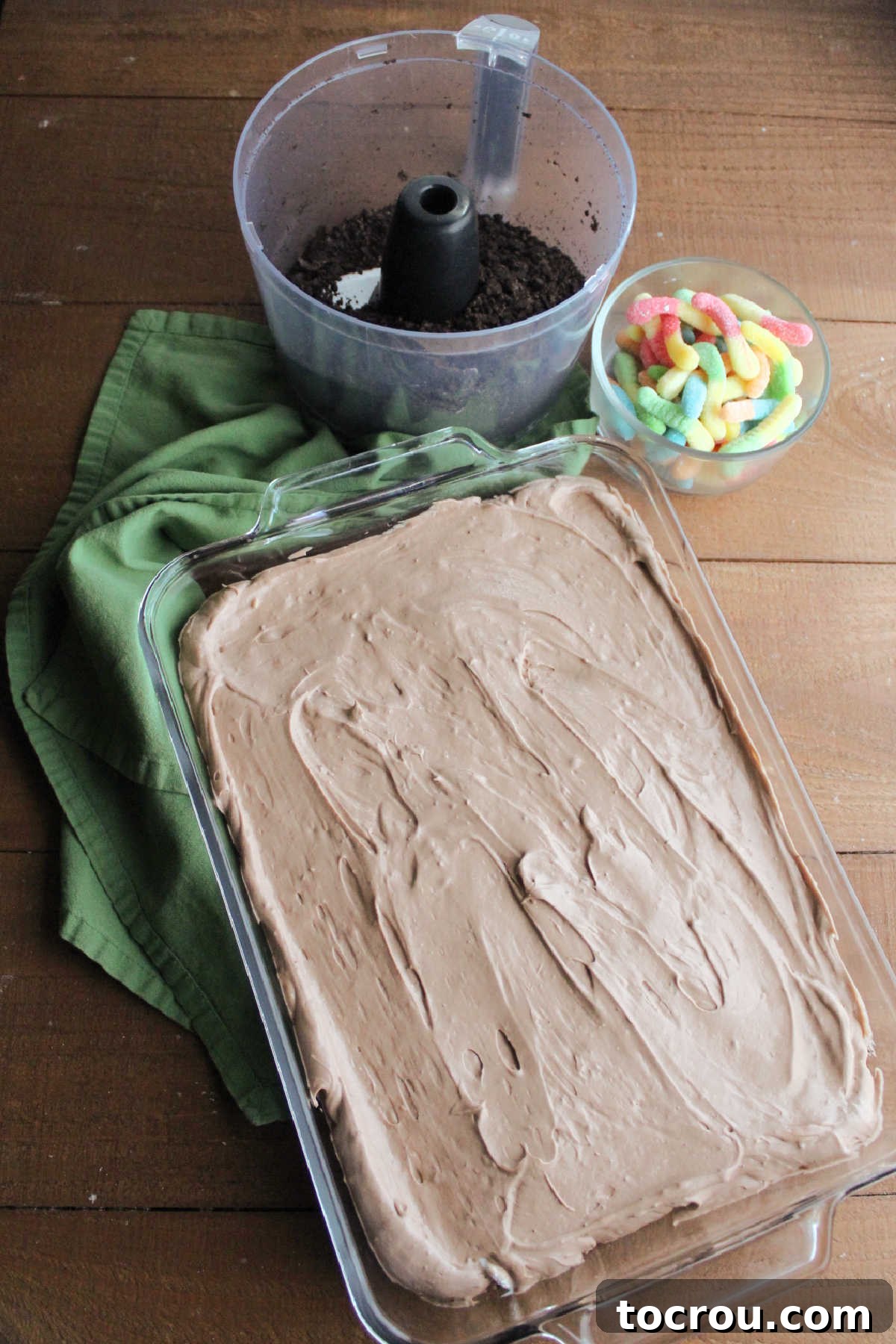 Chocolate pudding and cream cheese mixture spread over bottom layer of Oreo crumbs in pan with the remaining Oreo crumbs and gummy worms in the background.