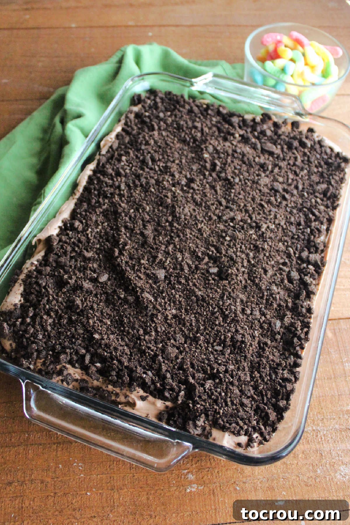 Dirt cake topped with Oreo crumbs with sour gummy worms nearby, ready to go on the top to finish the dessert.