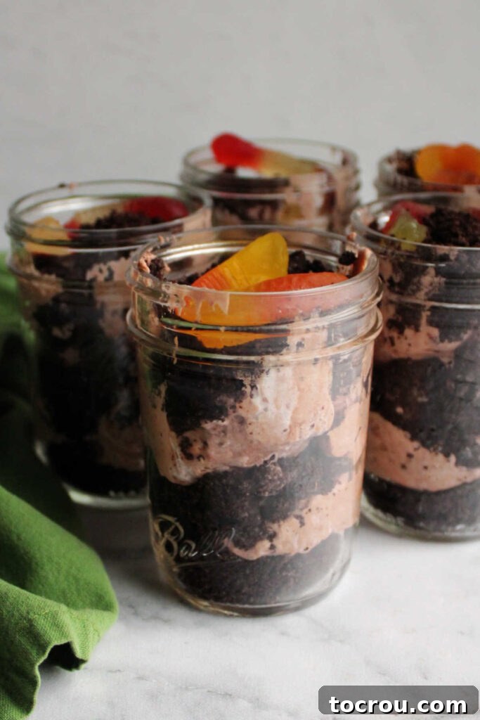 Jars of dirt pudding with layers of Oreo crumbs, chocolate pudding mixture and a gummy worm on top.