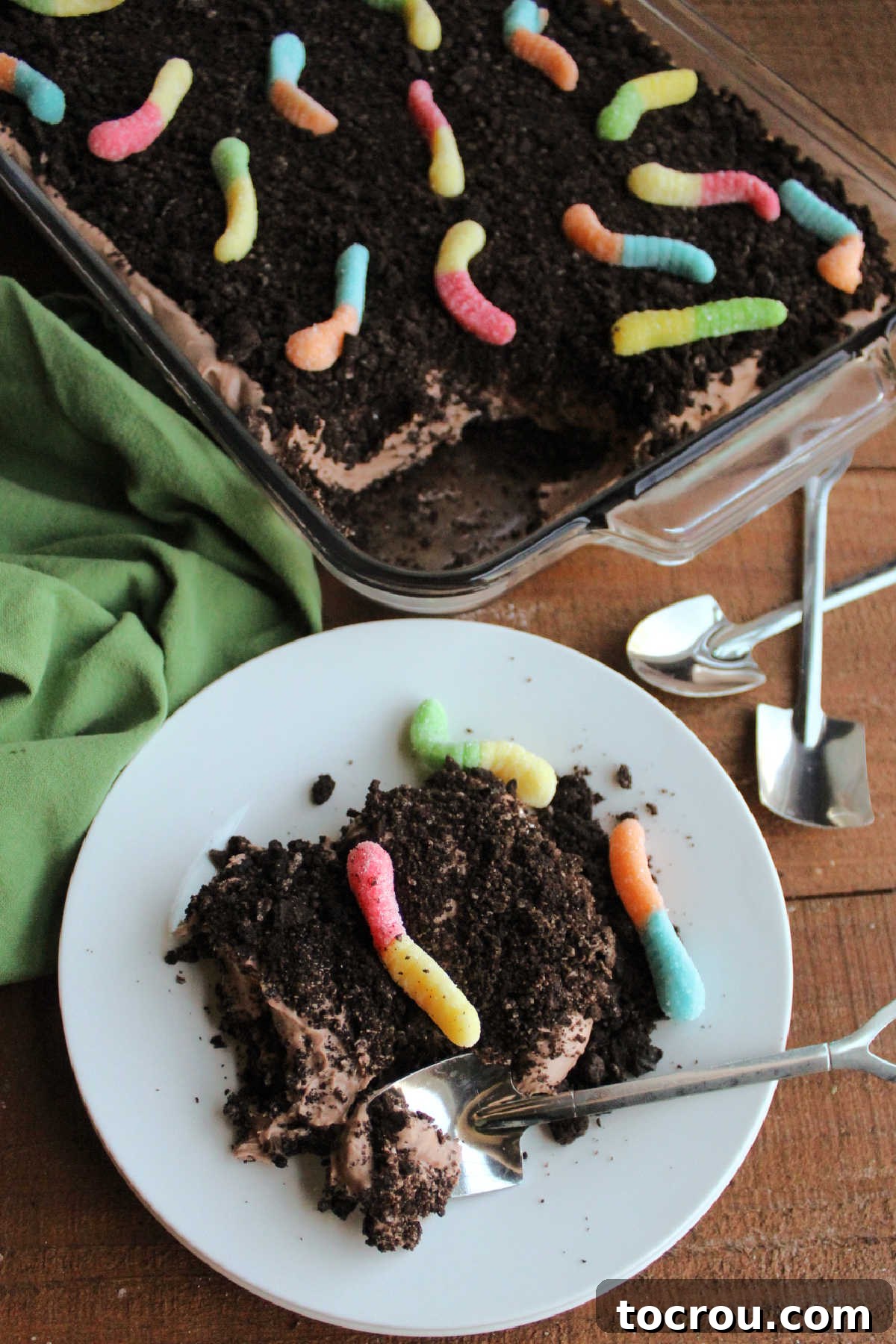 Serving of dirt cake on small plate showing fluffy chocolate pudding center, Oreo crumbs and gummy worms served with a shovel shaped spoon, ready to eat.