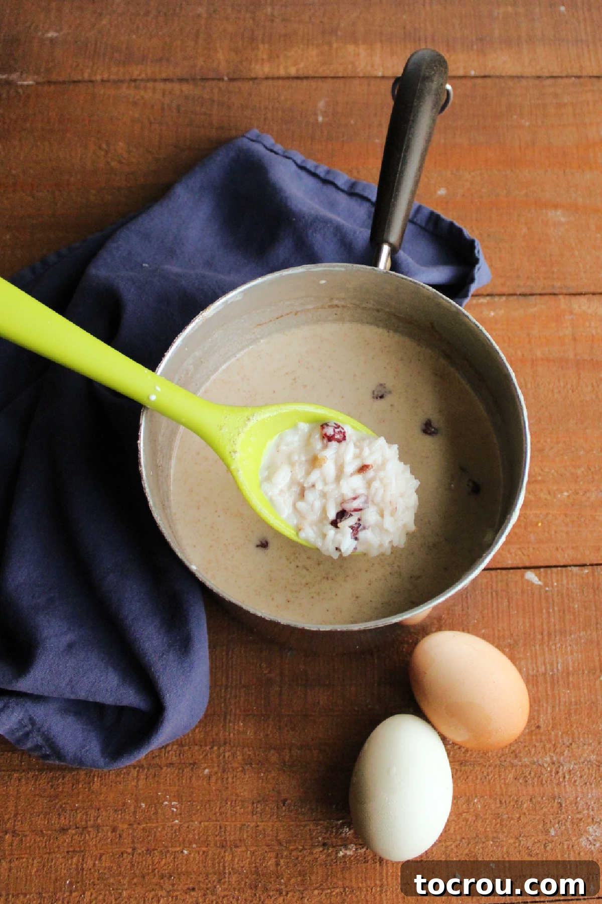 Mixing Rice Pudding Base Saucepan with cooked rice mixed with evaporated milk, craisins, sugar, vanilla and spices next to eggs.