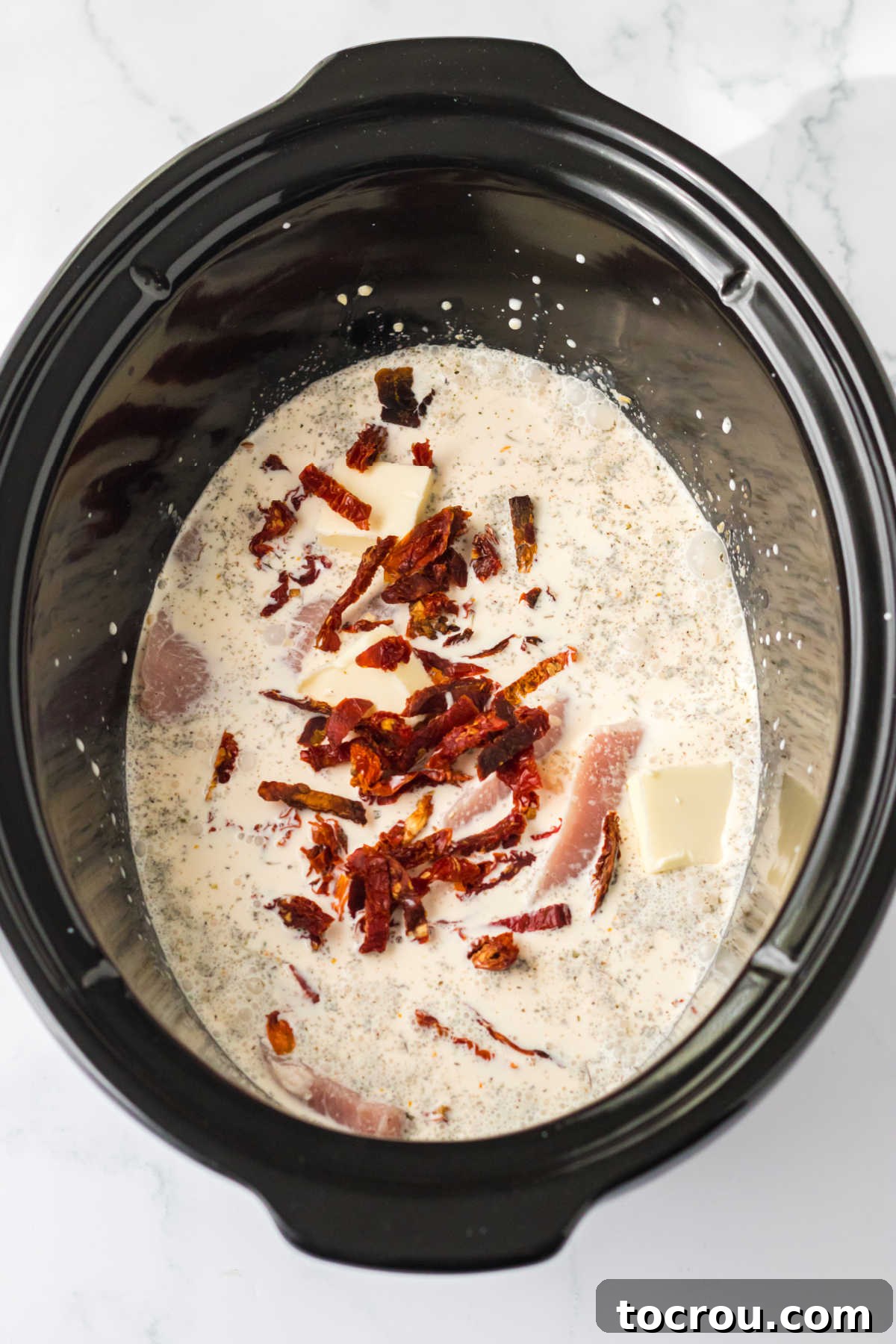 Pork chops in slow cooker with cream and broth mixture, topped with sun dried tomatoes and pats of butter, ready to cook.