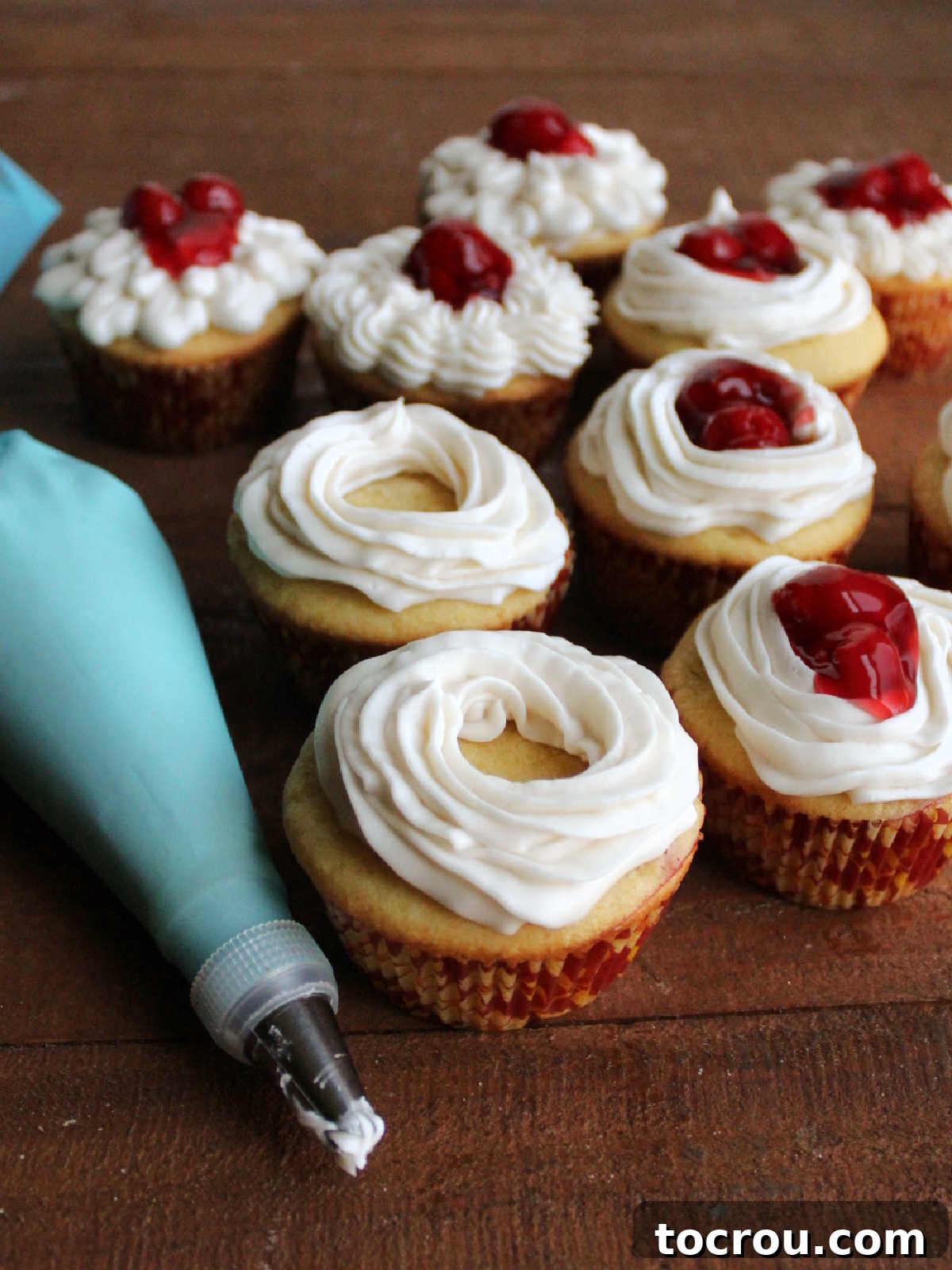 Piping bag with vanilla buttercream next to cupcakes with buttercream rings piped on top, some filled with cherry pie filling.