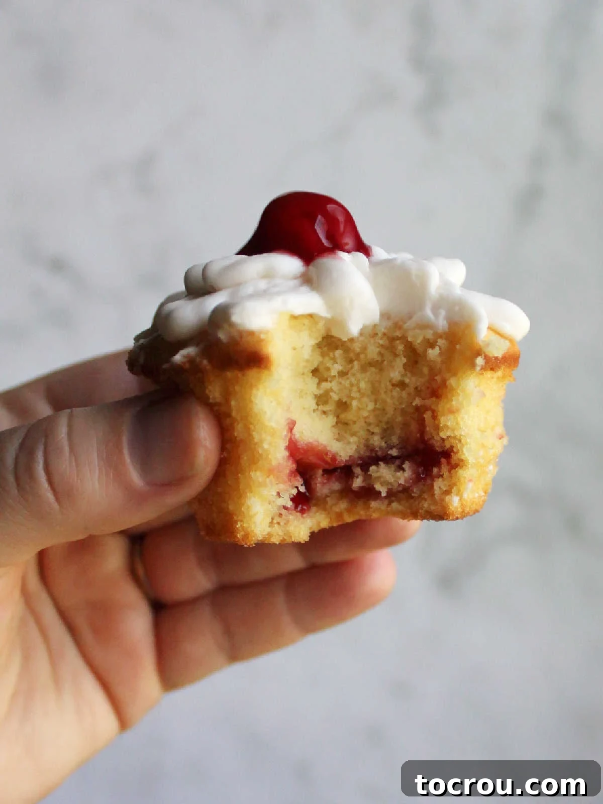 Hand holding cupcake with a bite missing, showing layer of cherry pie filling baked inside and frosting and more cherries on top.