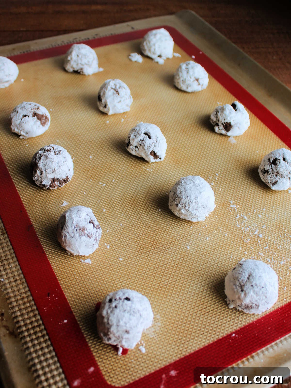 Scoops of chocolate gooey butter cookie dough coated in powdered sugar on baking sheet, ready to go in the oven.