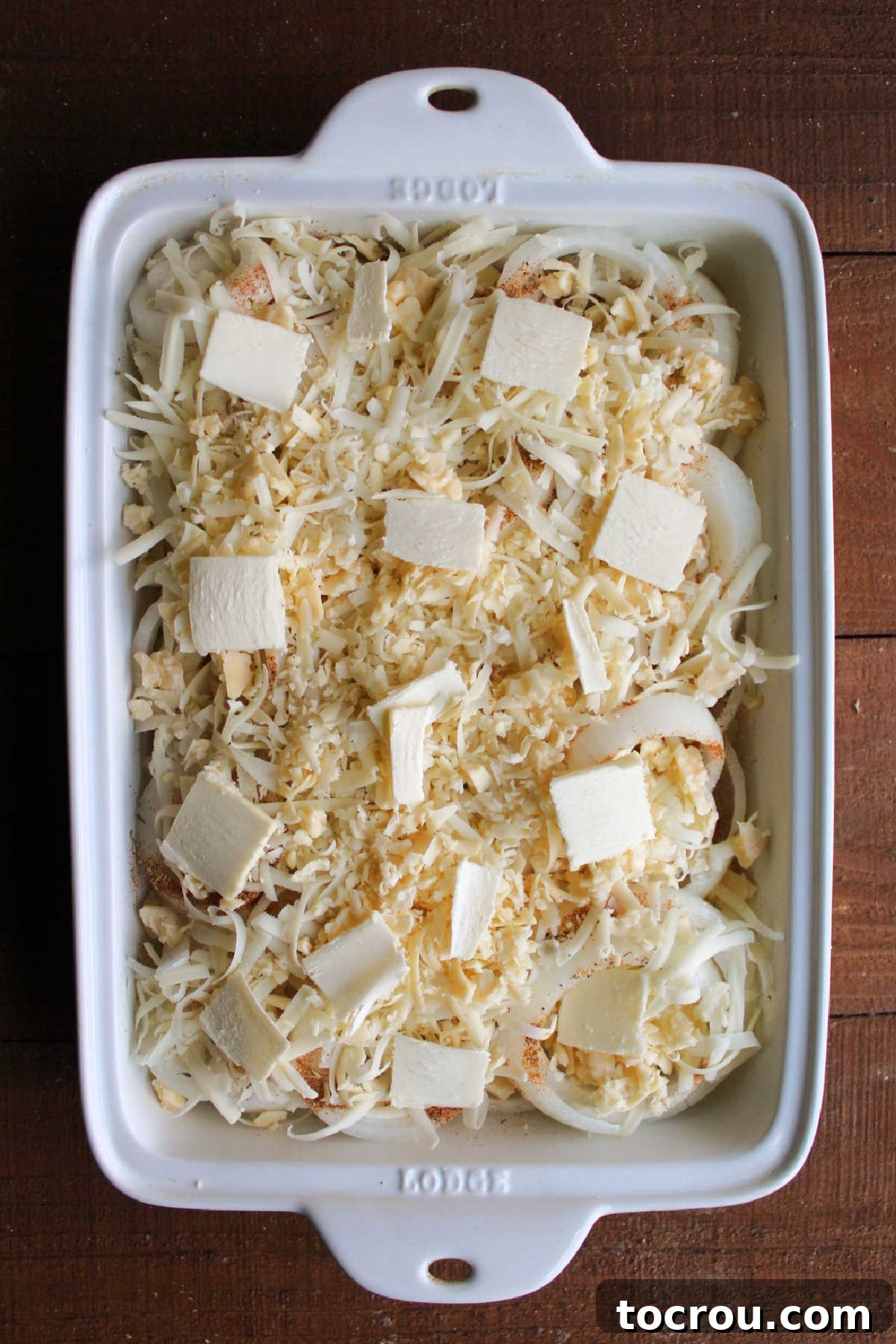Shredded cheese and pats of butter over onions in baking pan, ready to be covered with foil and baked.