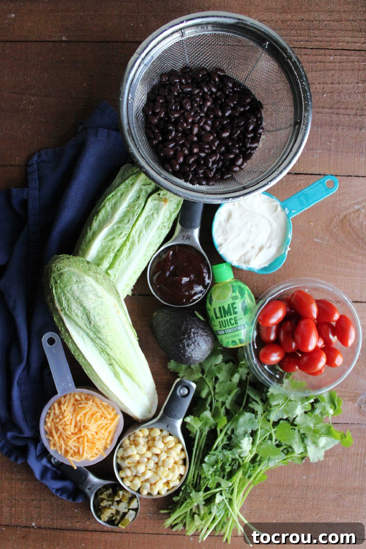 Ingredients including romaine lettuce, black beans, cherry tomatoes, cilantro, corn, cheese, jalapenos, mayonnaise, bbq sauce, and lime juice ready to be made into cowboy salad.