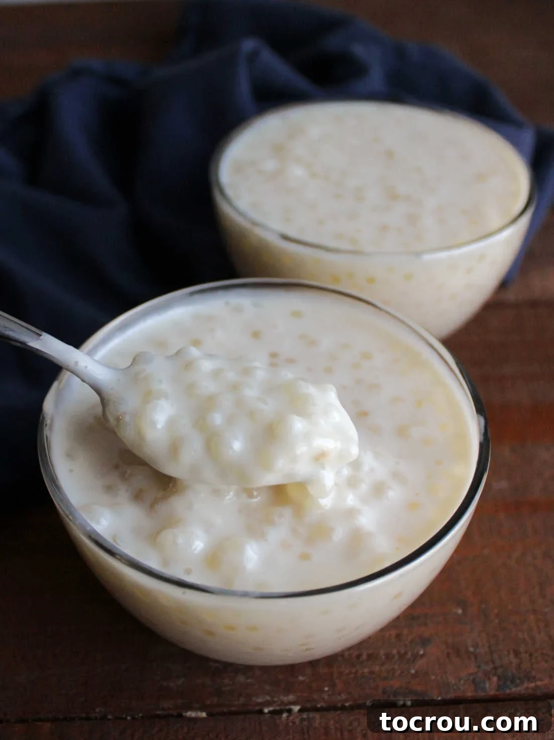 A spoon delicately scooping a mouthful of perfectly set tapioca pudding from a small glass bowl, highlighting its creamy texture and visible pearls.