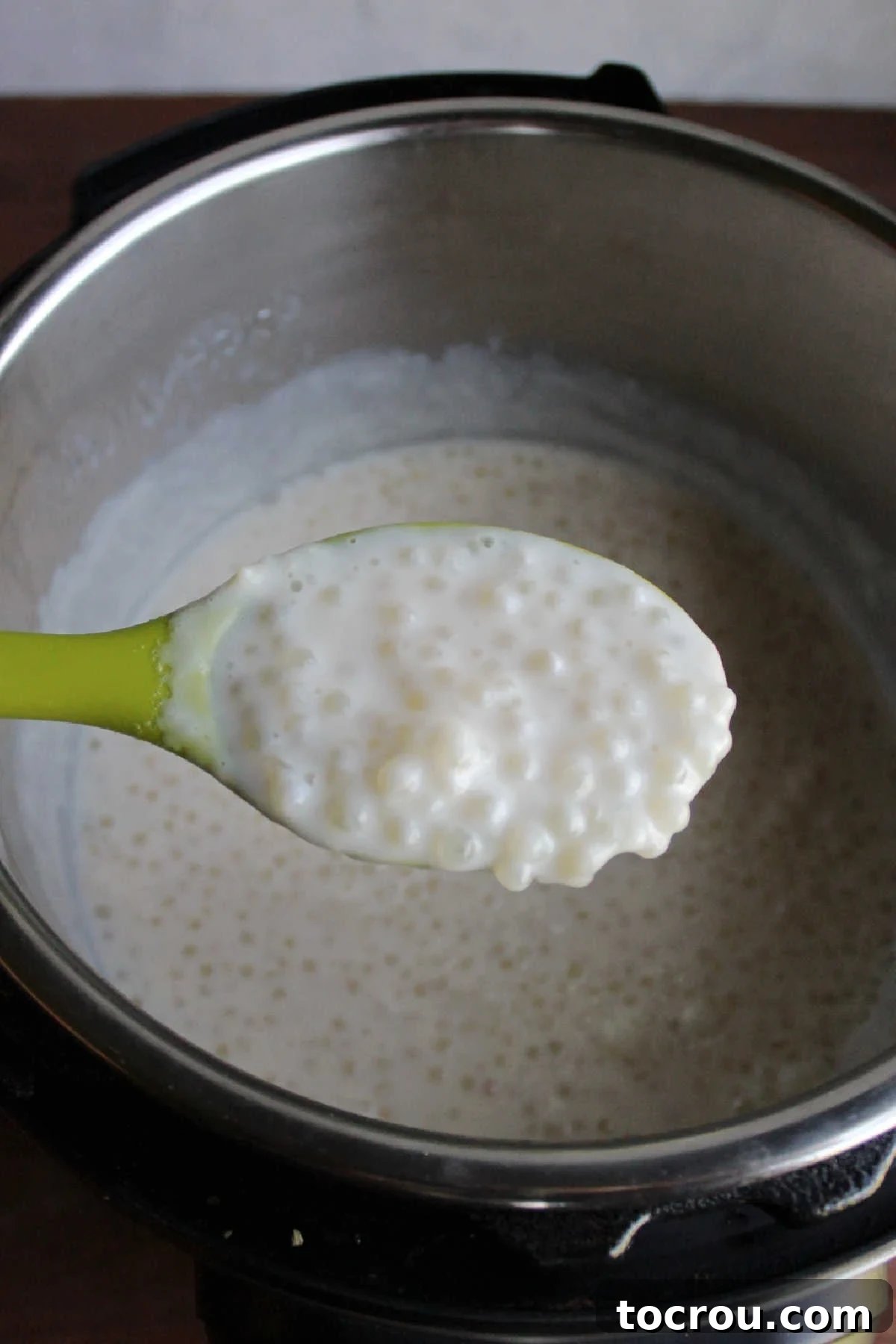 A large serving spoon holding a generous scoop of warm, fresh tapioca pudding, poised over the Instant Pot containing the remaining batch.