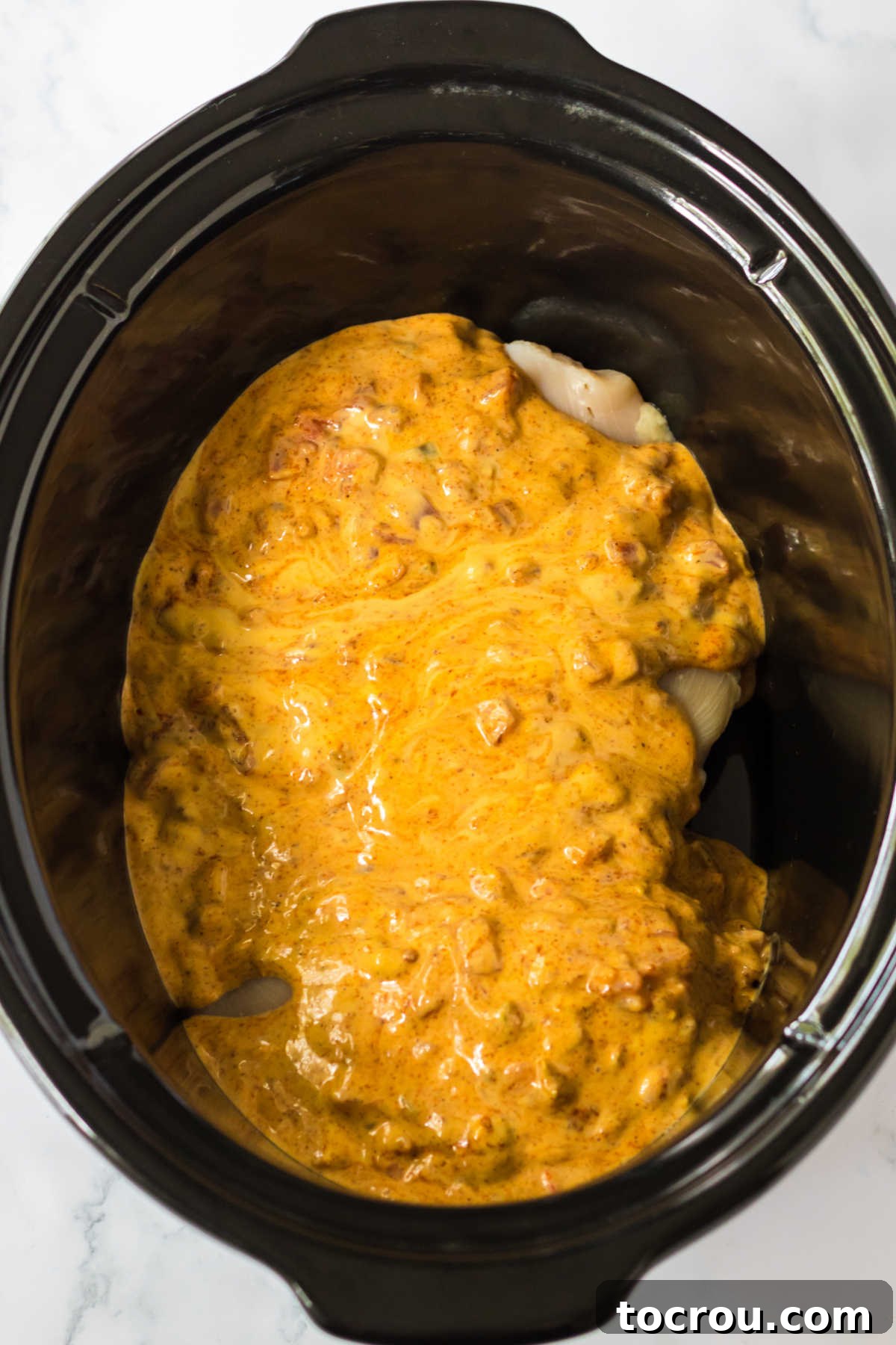 Chicken in Slow Cooker with Sauce The crockpot insert filled with raw chicken breasts, covered in a vibrant sauce made from queso, Rotel diced tomatoes with green chilies, and taco seasoning, poised for cooking.