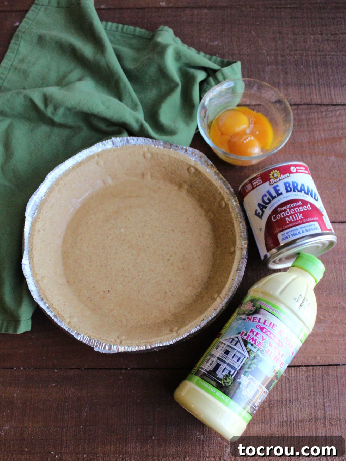 Essential ingredients for a simple key lime pie: a bottle of key lime juice, a can of sweetened condensed milk, separated egg yolks, and a ready-made graham cracker pie crust.