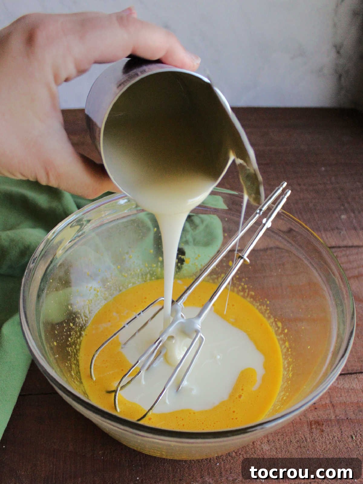A stream of rich, white sweetened condensed milk being poured into a bowl containing vibrant yellow beaten egg yolks, illustrating a key step in preparing the pie filling.