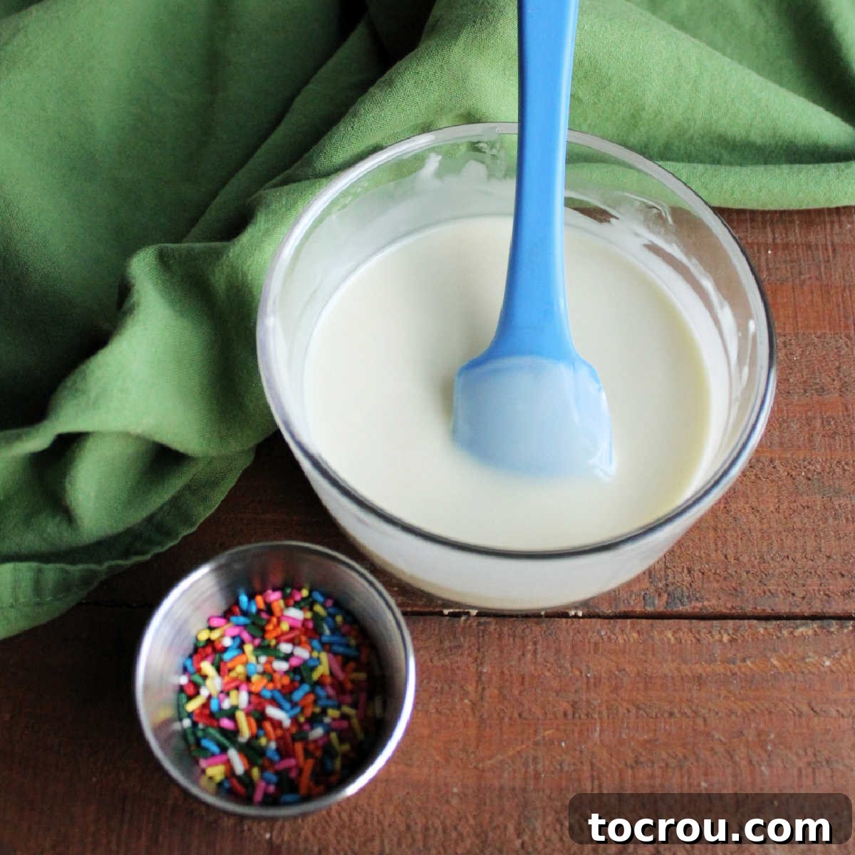 DIY Unicorn Rainbow Glaze 6 Melted white chocolate and coconut oil in small glass bowl next to a container of colorful sprinkles.