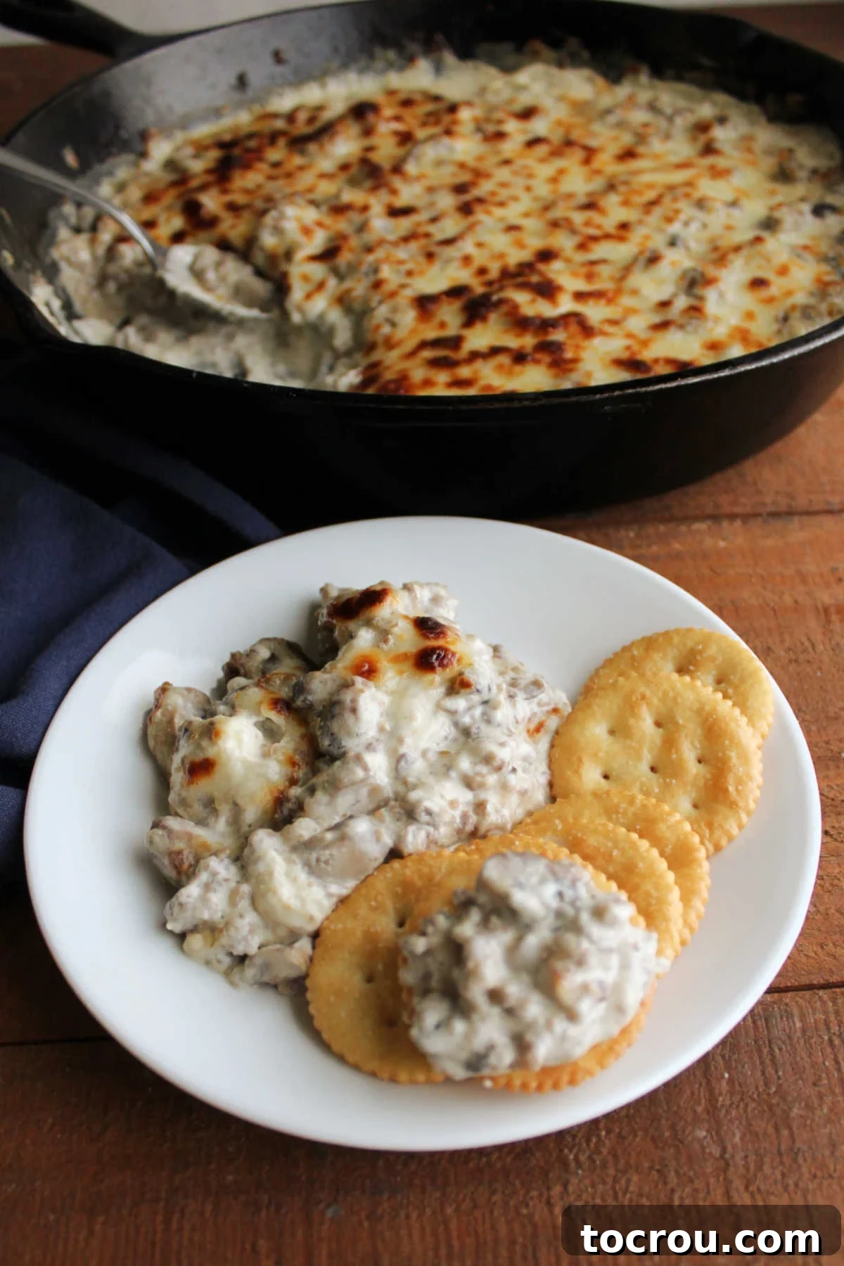 Creamy Baked Sausage Mushroom Dip 3 A small white bowl of stuffed mushroom dip sits on a rustic wooden board, surrounded by golden crackers ready for dipping.