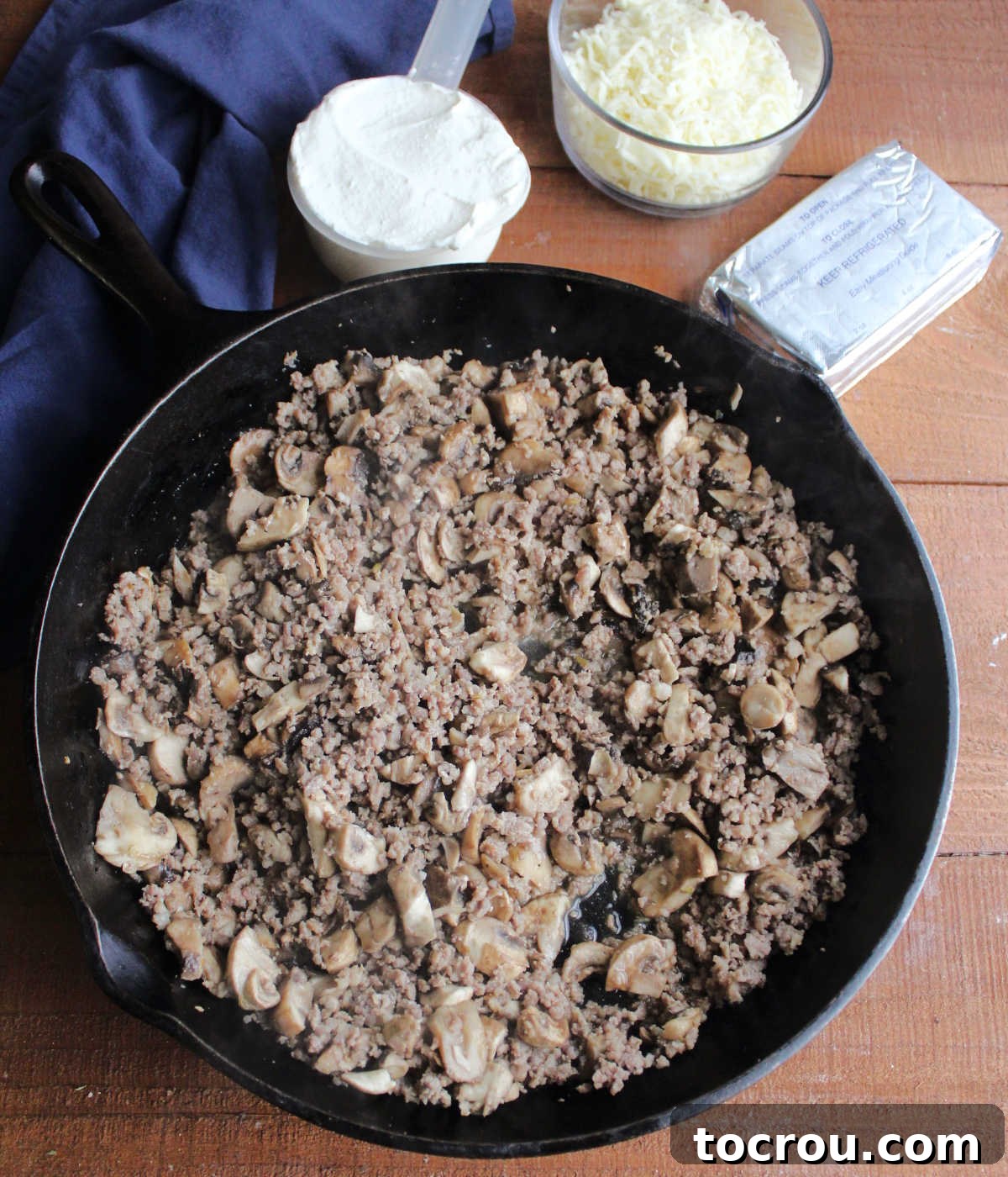Creamy Baked Sausage Mushroom Dip 5 A large cast iron skillet filled with browned sausage and cooked mushrooms, surrounded by bowls of cream cheese, sour cream, and shredded mozzarella cheese ready to be mixed in.