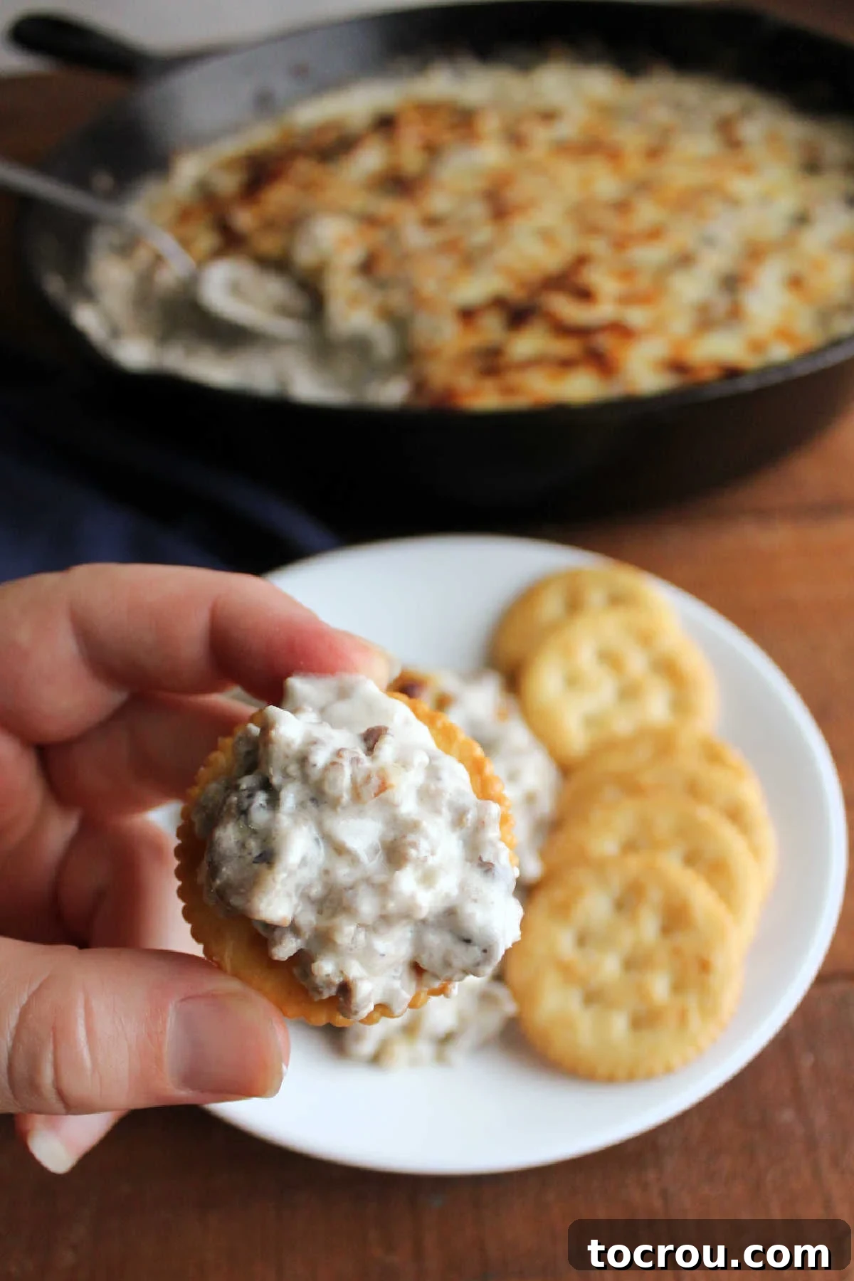 Creamy Baked Sausage Mushroom Dip 9 A hand holds a cracker piled high with creamy, cheesy stuffed mushroom dip, showcasing visible bits of sausage and mushroom.