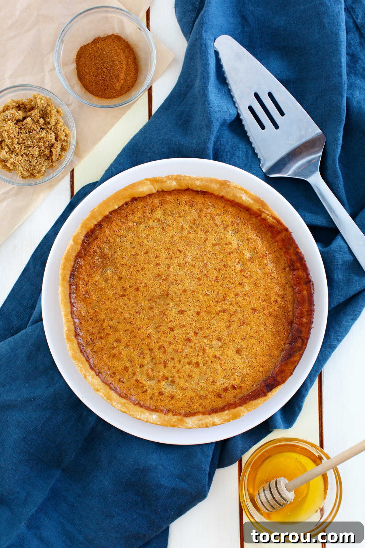 Enjoy this honey sweetened twist on a chess pie. With a kiss of lemon, honey richness and lots of chess pie goodness, it is a perfect treat for almost any occasion! whole honey chess pie with bowls of honey, cinnamon and brown sugar nearby and pie serve ready to go.