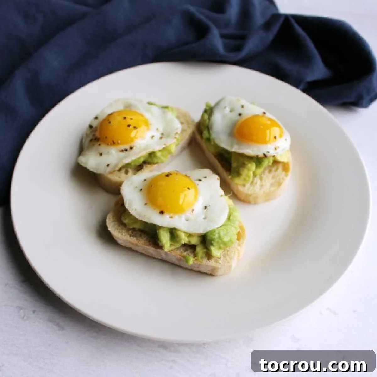 Three avocado and quail egg crostini elegantly arranged on a small white plate, ready to serve as a gourmet appetizer or brunch item.