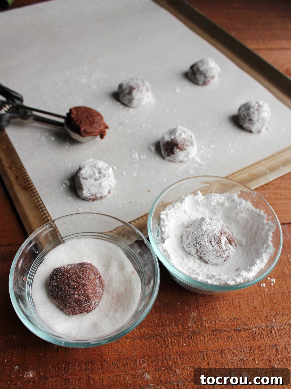 Rolling balls of soft chocolate cookie dough in granulated sugar then powdered sugar and putting them on a parchment lined baking tray.