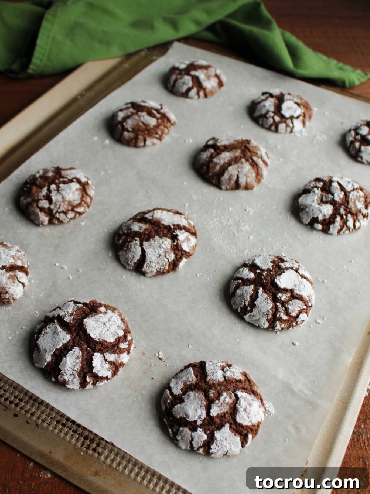 Freshly baked chocolate crinkle cookies on parchment lined baking tray showing crackled powdered sugar exterior.