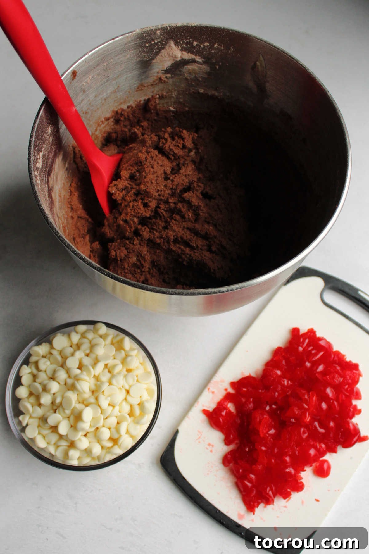 Ingredients for chocolate cherry chip cookies prepped for mixing. Mixing bowl filled with rich chocolate cookie dough, alongside bowls of chopped maraschino cherries and creamy white chocolate chips, ready to be combined.