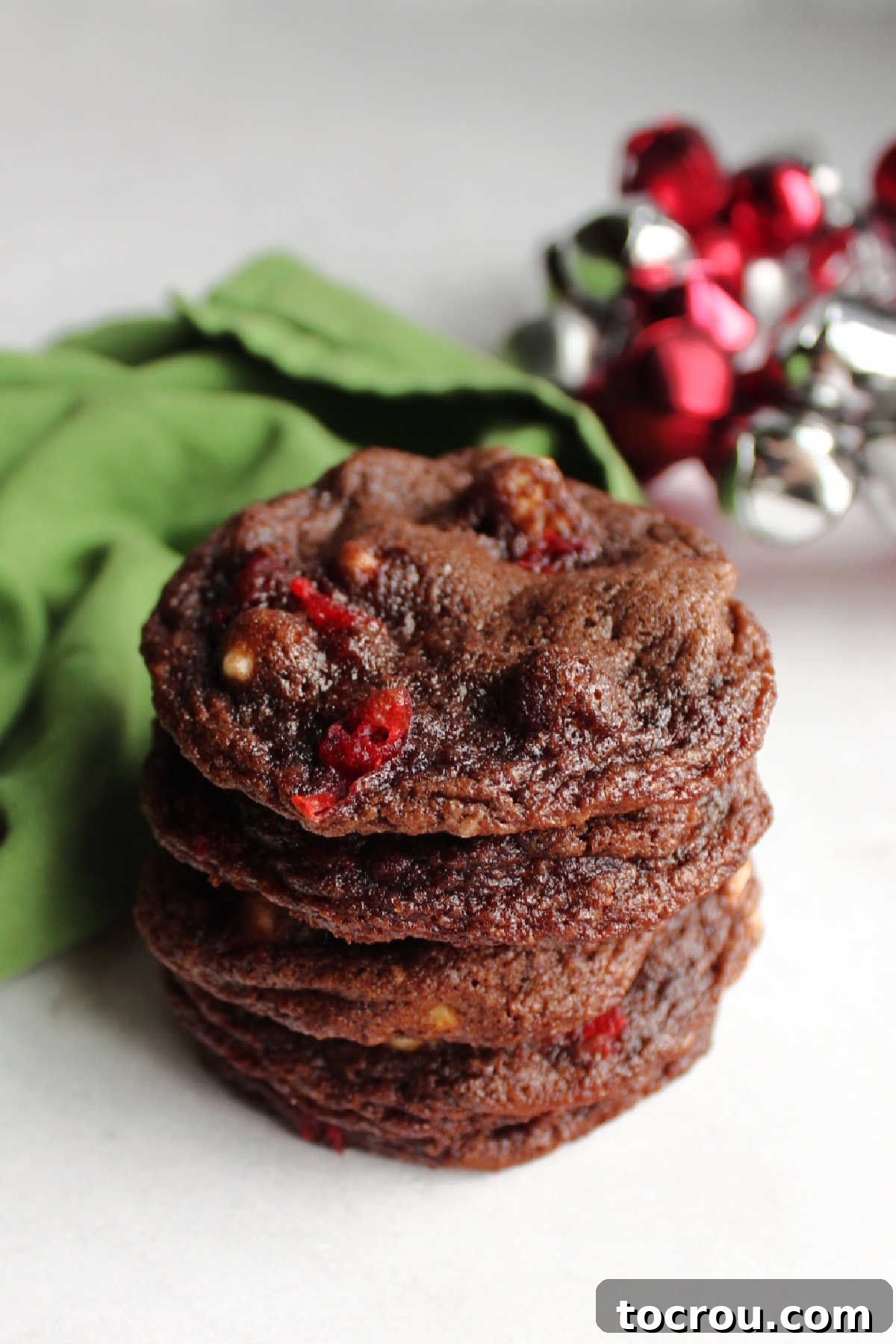 These cookies are rich, chocolaty, and incredibly delicious, dotted with cherries and white chocolate chips for that beloved chocolate-covered candy experience. A charming stack of chocolate cherry chip cookies with festive holiday bells in the blurred background, evoking a cozy Christmastime atmosphere.