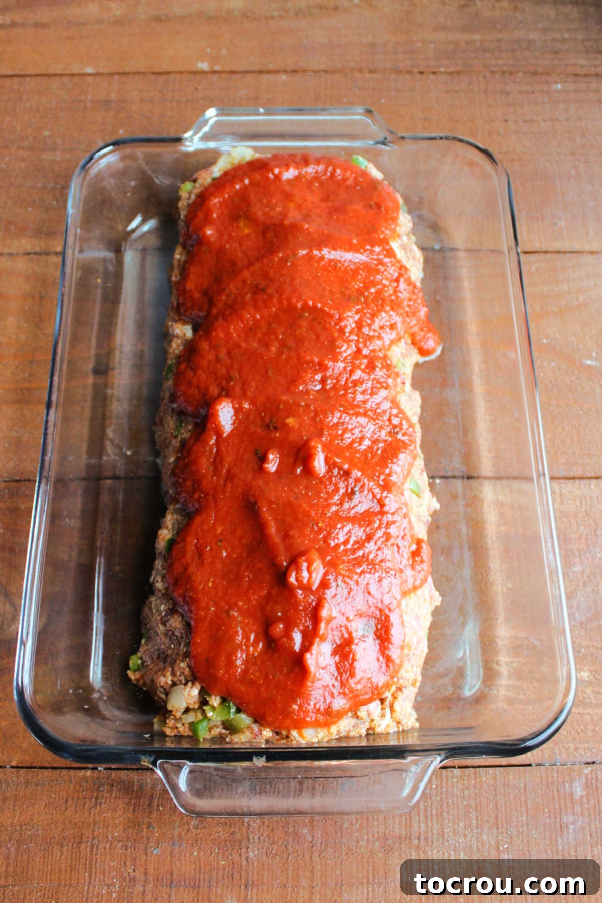 Marinara sauce spread evenly on top of raw Italian sausage meatloaf in a baking pan, ready to be placed into the oven for baking.