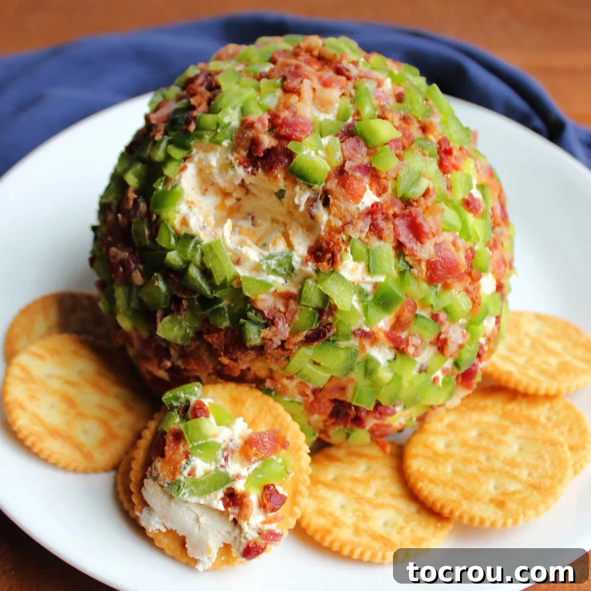 Zingy Bacon Jalapeno Cheese Ball 2 Cheese ball coated in bits of jalapeno pepper and bacon with a little bit missing showing cheesy inside on plate with crackers, ready to eat.