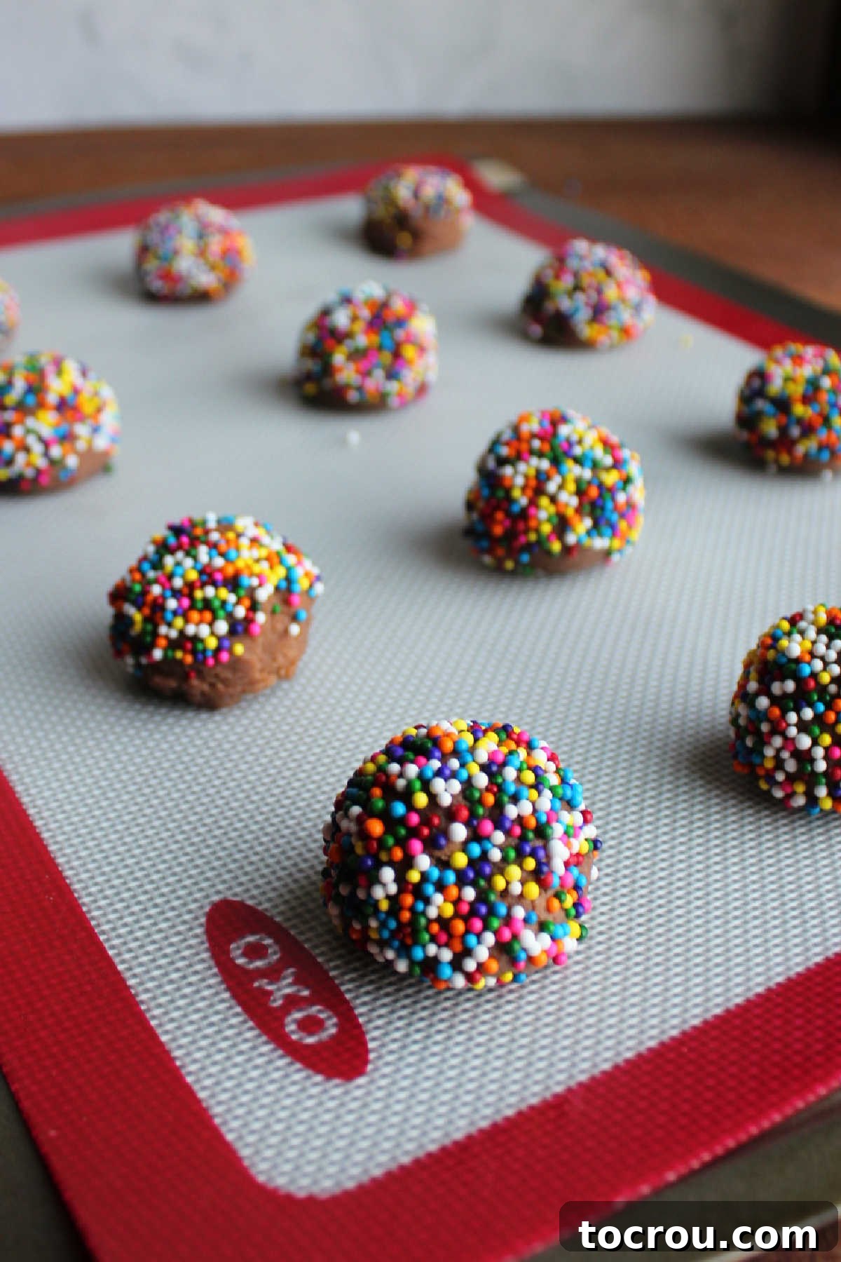 Scoops of chocolate cookie dough, some rolled in colorful sprinkles, on a silicone baking mat lined pan.