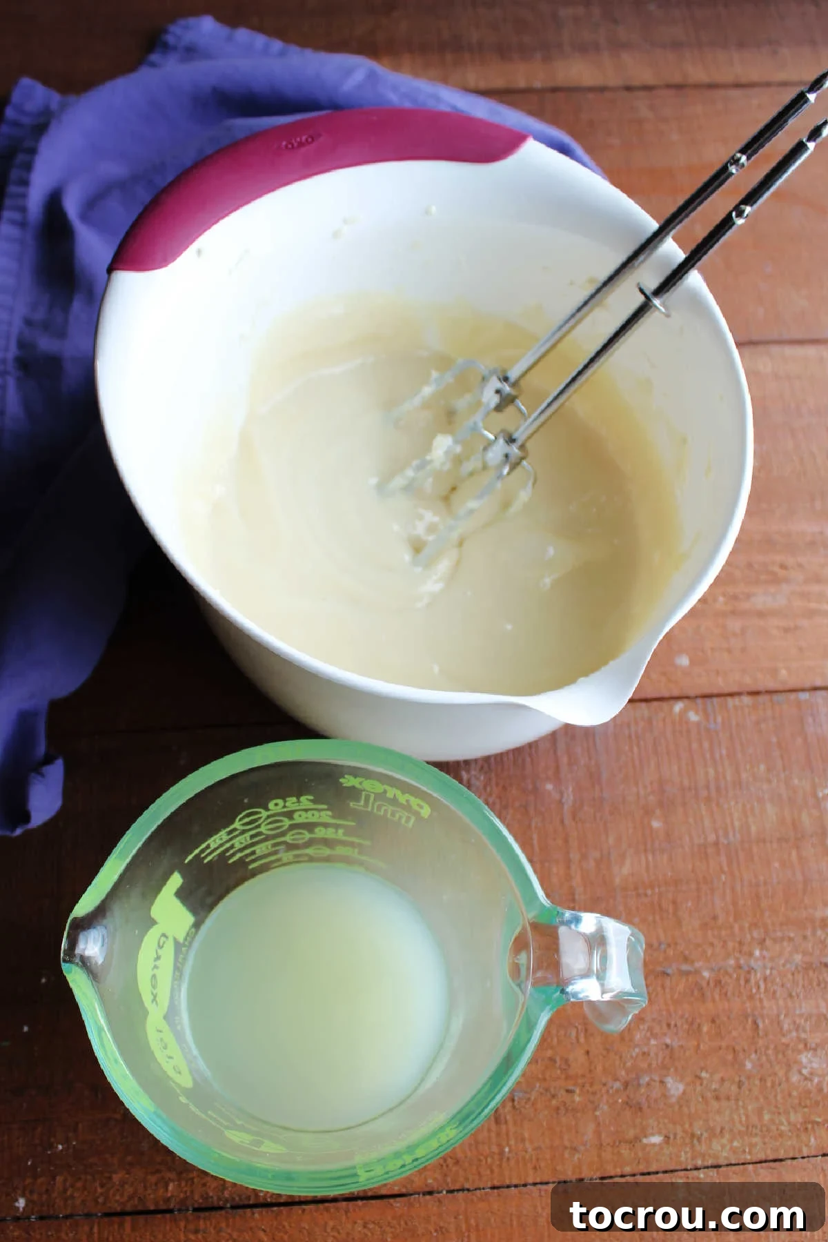 Mixing bowl containing smooth cream cheese and condensed milk mixture next to a measuring cup with lemon juice inside.