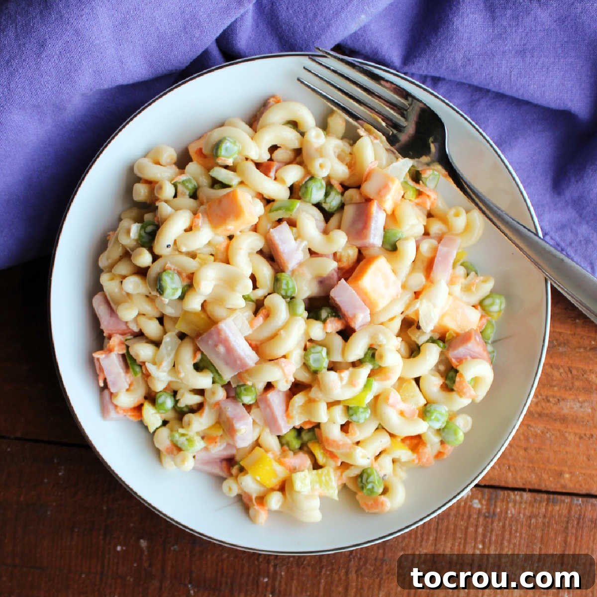 Lunch size portion of sweet pasta salad with peas, ham, cheese, onions, carrots, and peppers in a creamy tangy and sweet dressing with a fork, ready to eat.