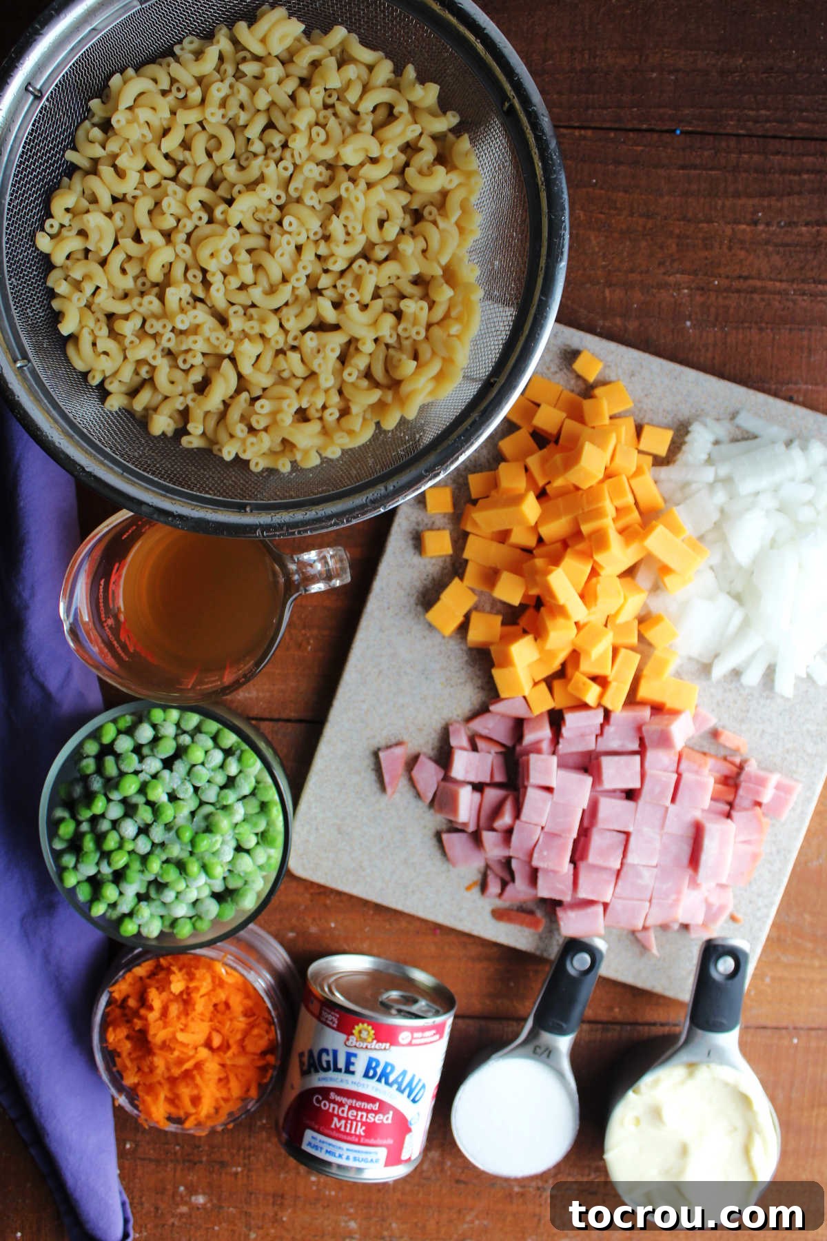Ingredients including cooked macaroni noodles, peas, vinegar, shredded carrots, sweetened condensed milk, sugar, mayonnaise, ham, cubed cheddar cheese, and diced onion ready to be made into sweet pasta salad.