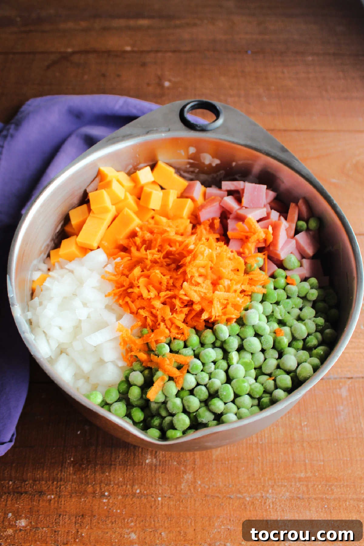 Frozen peas, shredded carrots, cubed ham and cheddar, and diced onion on top of pasta and dressing ready to be mixed in.