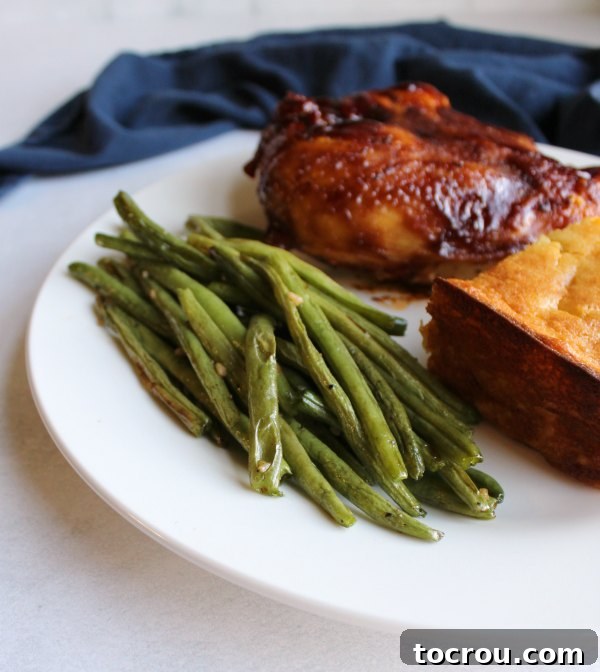 A dinner plate featuring a generous serving of roasted green beans alongside baked chicken and creamy corn pudding.